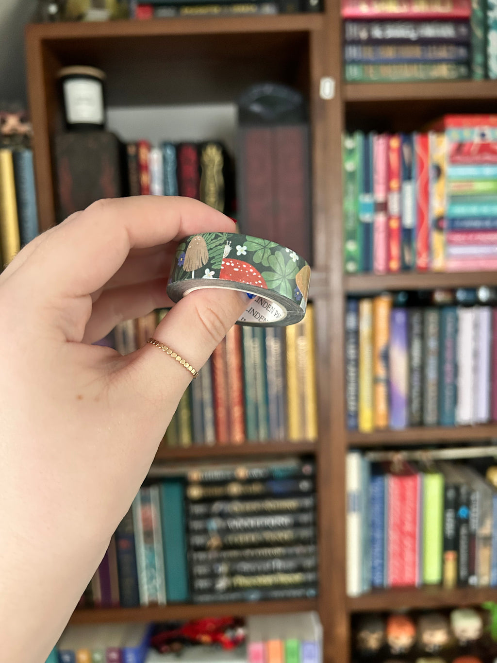 A hand holding a roll of decorative washi tape with a mushroom pattern, set against a blurred background of a bookshelf.