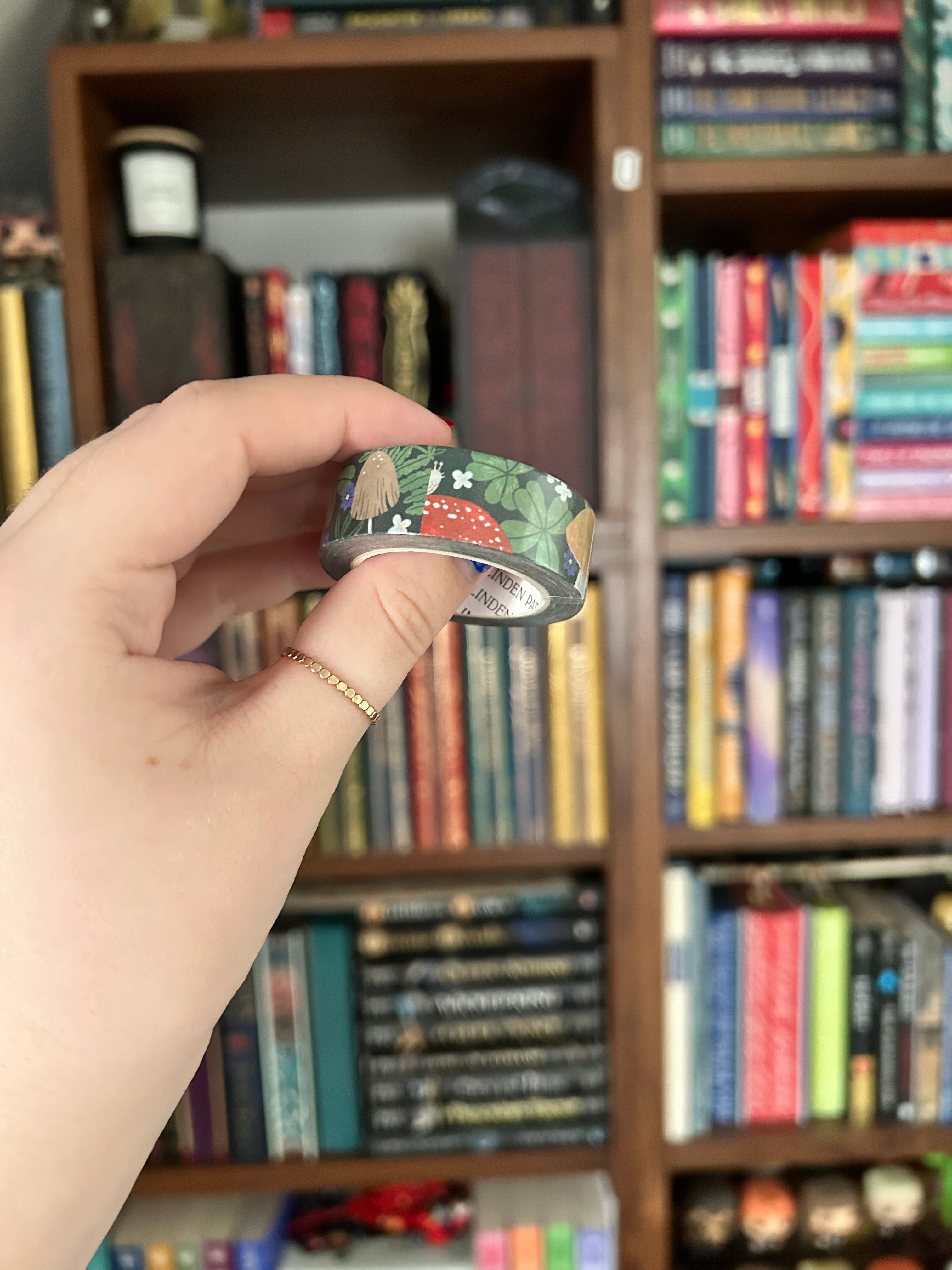 A hand holding a roll of decorative washi tape with a mushroom pattern, set against a blurred background of a bookshelf.