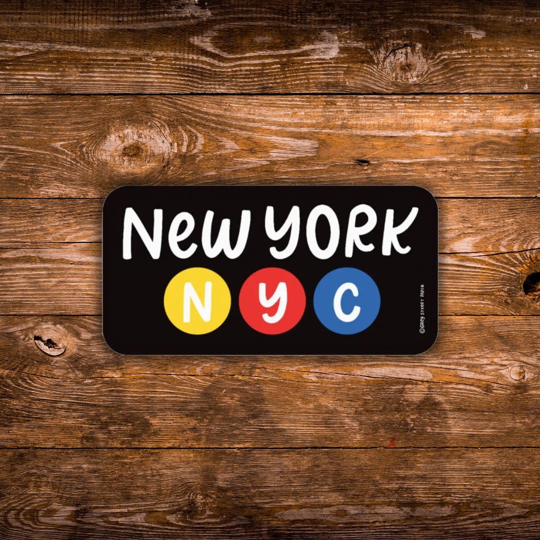 A rectangular black sign with rounded corners featuring the words "New York" in white above three colored circles labeled "N", "Y", and "C" in white. The sign is photographed against a rustic brown wooden plank background.