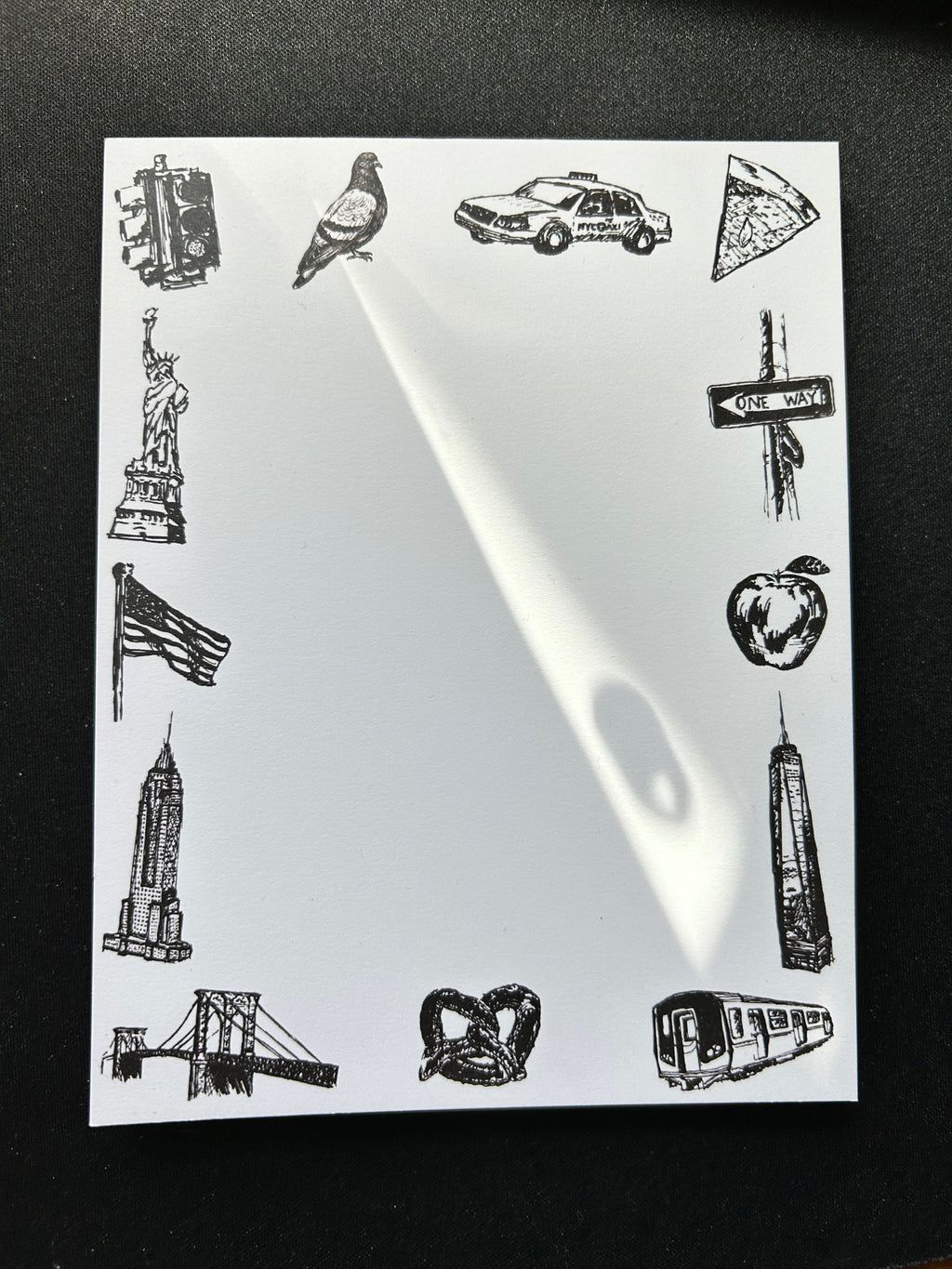 Illustrated New York City tear-away notepad featuring hand-drawn NYC landmarks and icons including the Statue of Liberty, Empire State Building, subway train, taxi, pizza, pigeon, and pretzel surrounding a blank writing area.