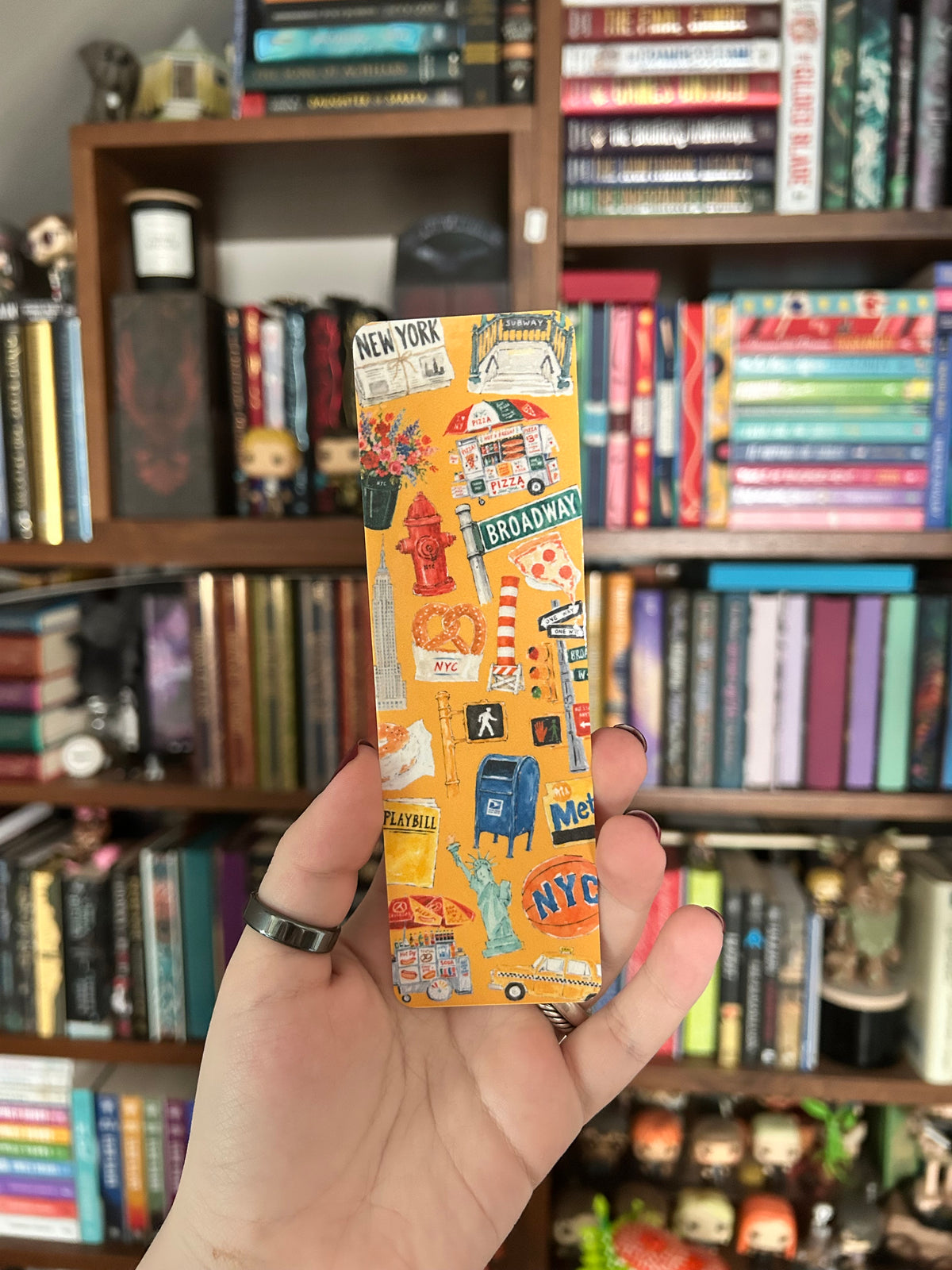 A hand holding a slim yellow bookmark decorated with colorful New York City illustrations, including street signs, a fire hydrant, a Statue of Liberty, and the words 'New York' and 'Broadway', in front of a blurred bookshelf background.