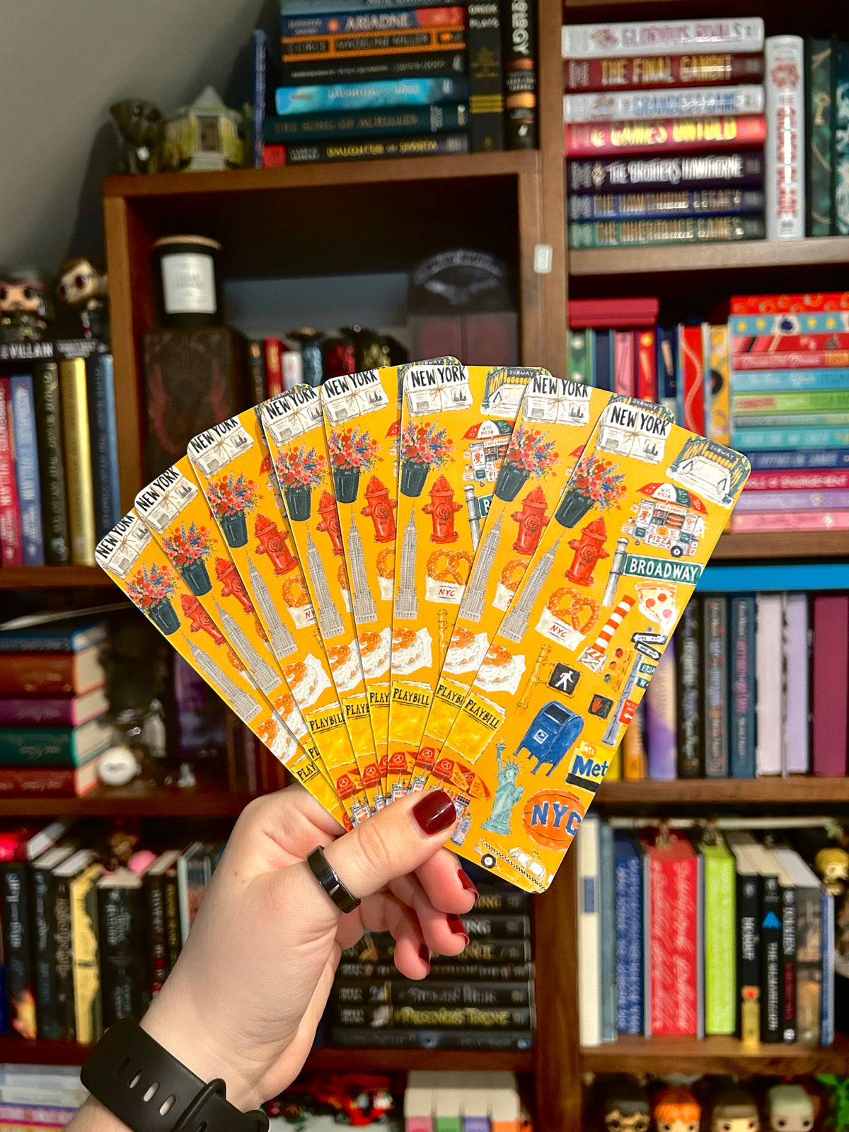 A hand holds a fan of yellow New York City themed bookmarks in front of a bookshelf filled with books.