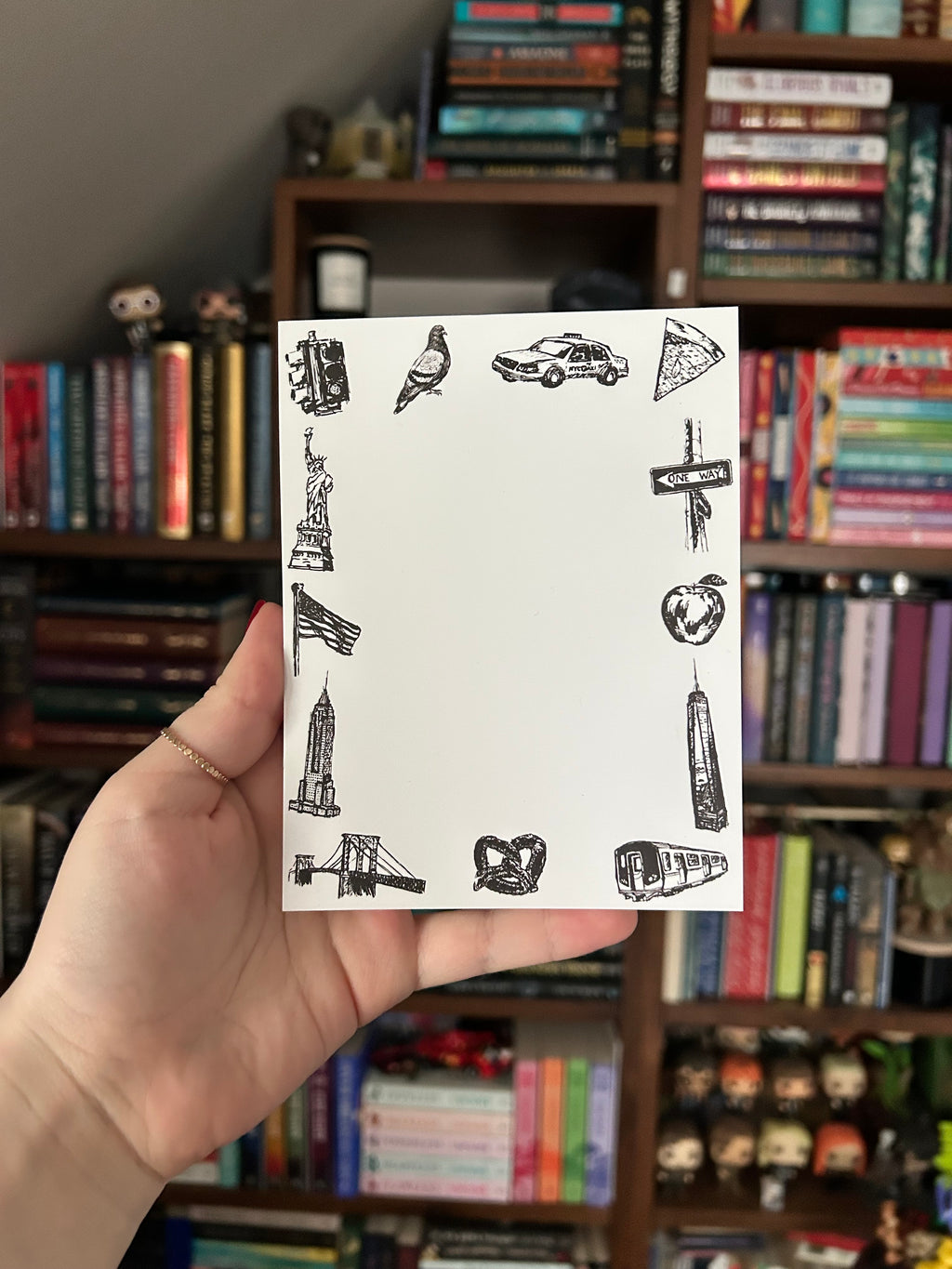 Illustrated New York City tear-away notepad featuring hand-drawn NYC landmarks and icons including the Statue of Liberty, Empire State Building, subway train, taxi, pizza, pigeon, and pretzel surrounding a blank writing area.