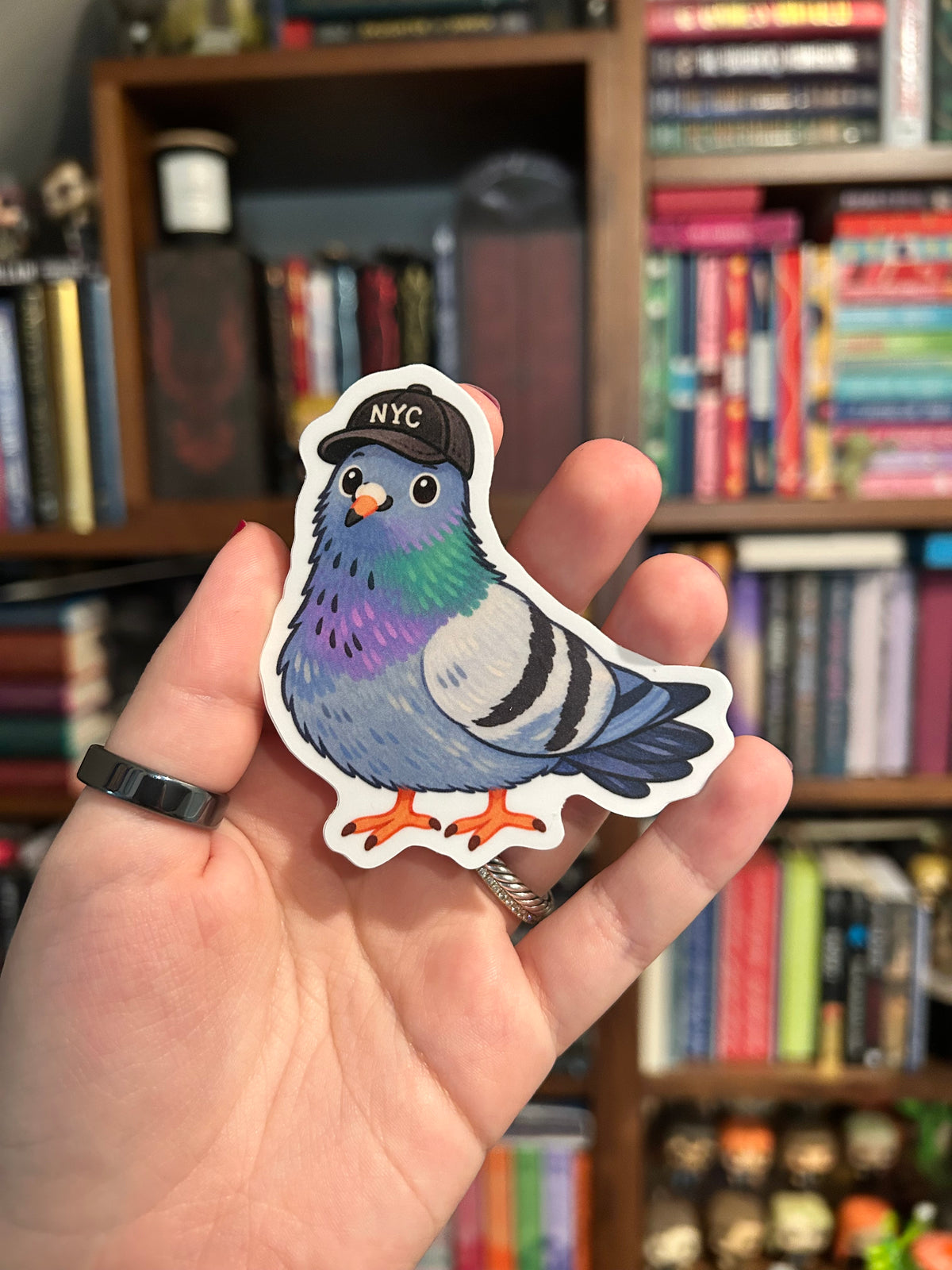 Illustrated blue-gray NYC pigeon sticker wearing a black New York City baseball cap, with iridescent green and purple neck feathers, orange feet, and a white border.