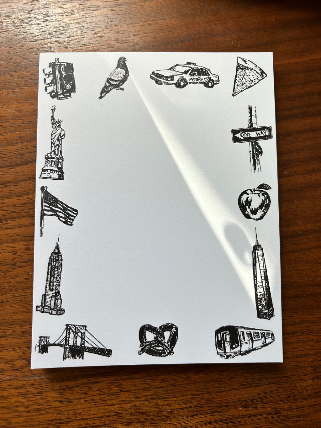 Illustrated New York City tear-away notepad featuring hand-drawn NYC landmarks and icons including the Statue of Liberty, Empire State Building, subway train, taxi, pizza, pigeon, and pretzel surrounding a blank writing area.