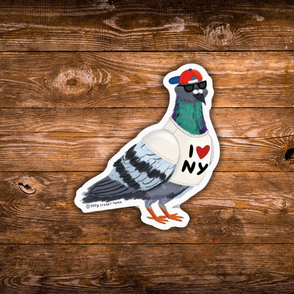 A die-cut sticker of a stylized pigeon wearing a red and blue cap, black sunglasses, and a white shirt with 'I [heart] NY' printed on it, placed on a rustic brown wooden surface.