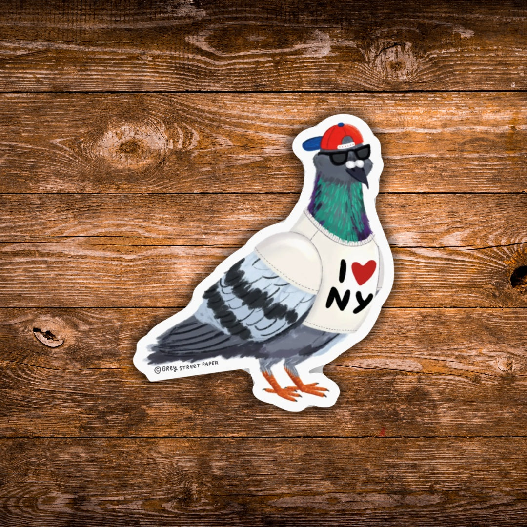 A die-cut sticker of a stylized pigeon wearing a red and blue cap, black sunglasses, and a white shirt with 'I [heart] NY' printed on it, placed on a rustic brown wooden surface.