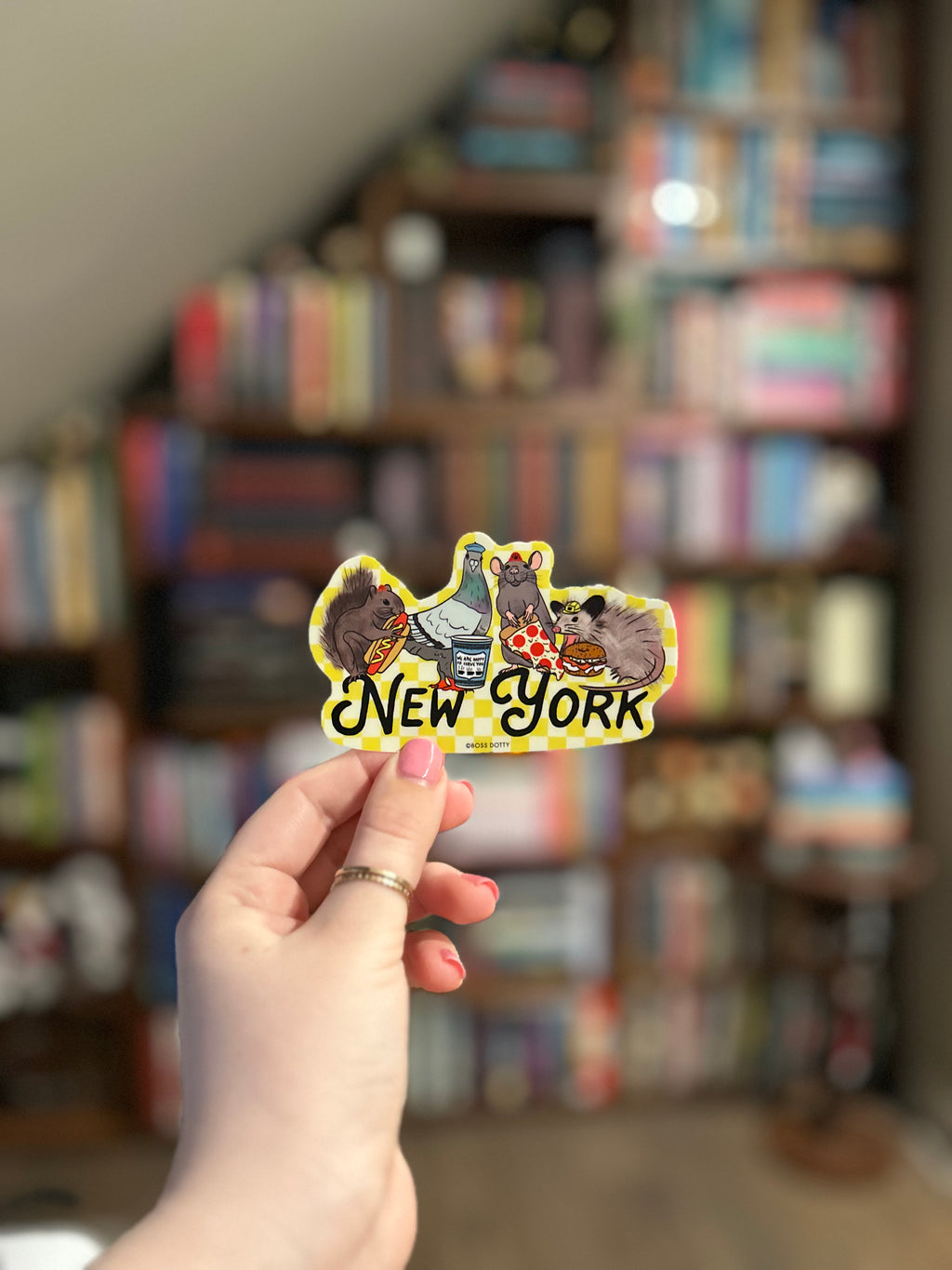 A hand holds a die-cut sticker featuring cartoon animals and the text 'New York' in front of a blurred bookshelf background.