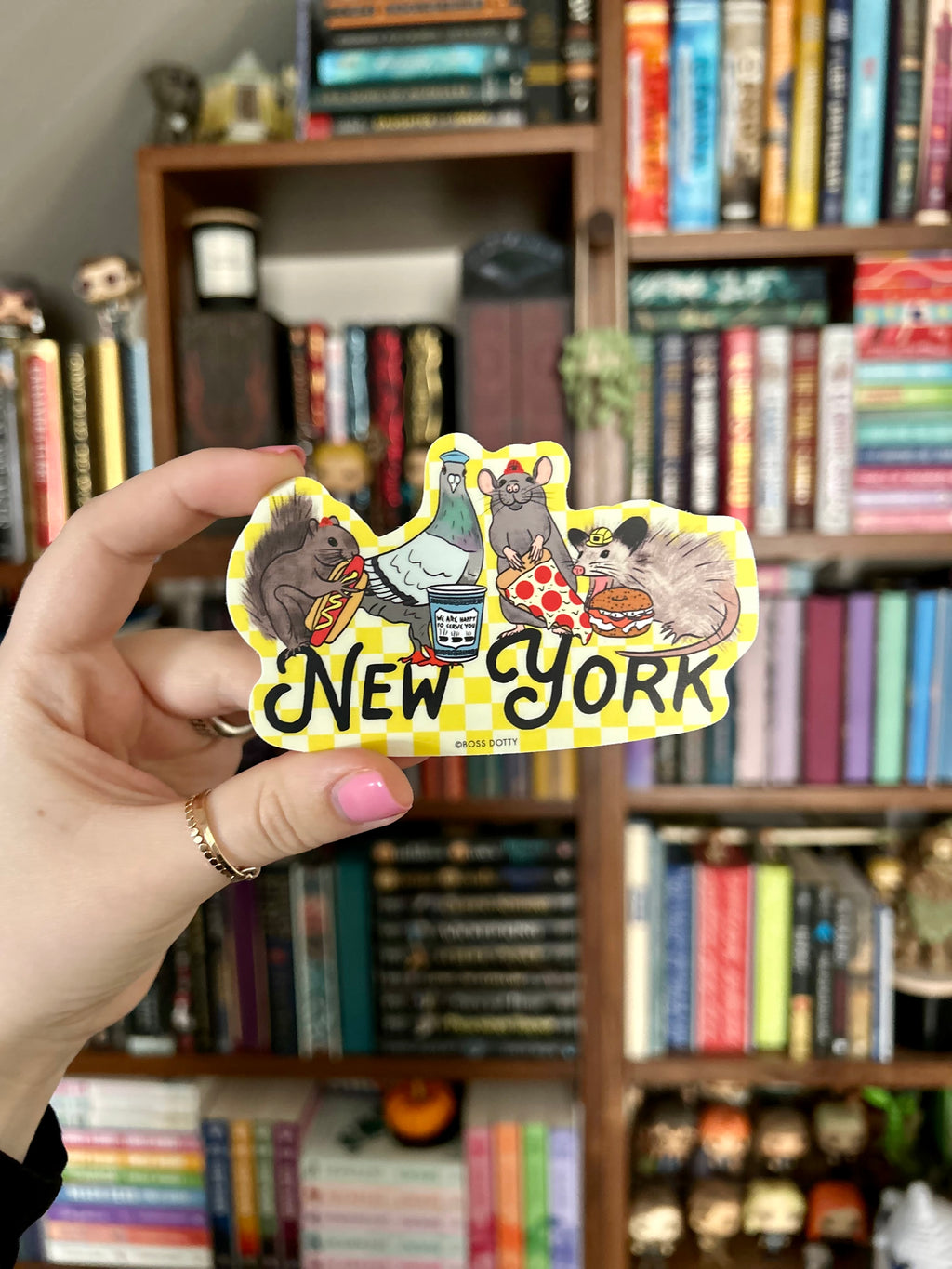 A hand holds a die-cut sticker featuring cartoon animals and the text 'New York' in front of a blurred bookshelf background.