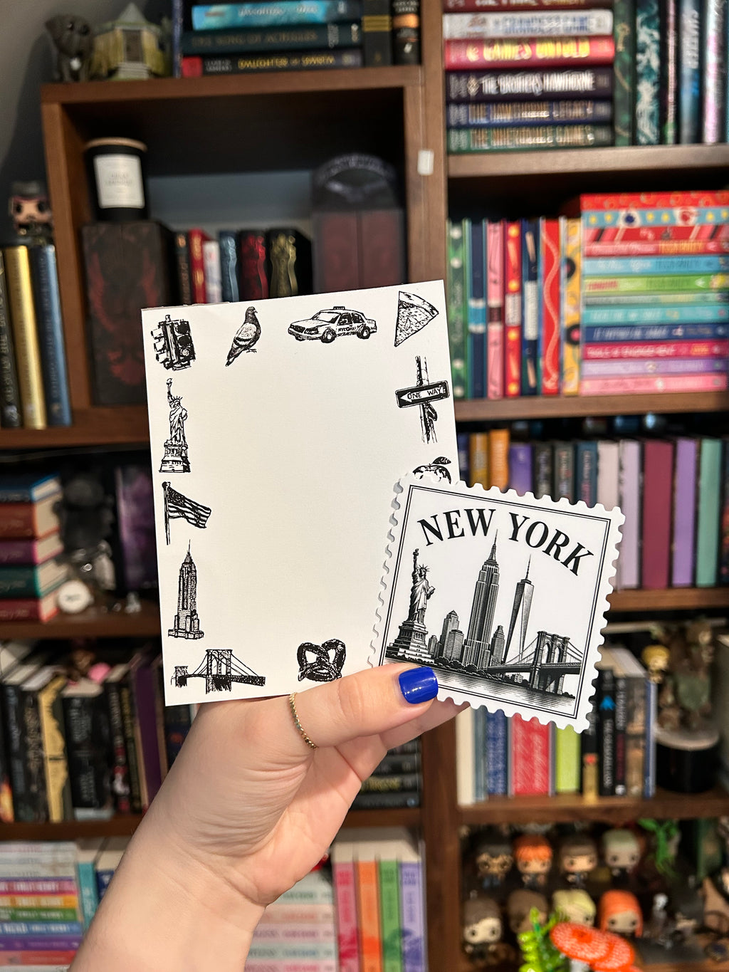 A hand with blue nail polish holds a white New York-themed notepad and sticker in front of a blurred bookshelf background. The sticker features a black-and-white New York skyline illustration and the words 'NEW YORK,' while the notepad has small black travel-themed doodles around the border.