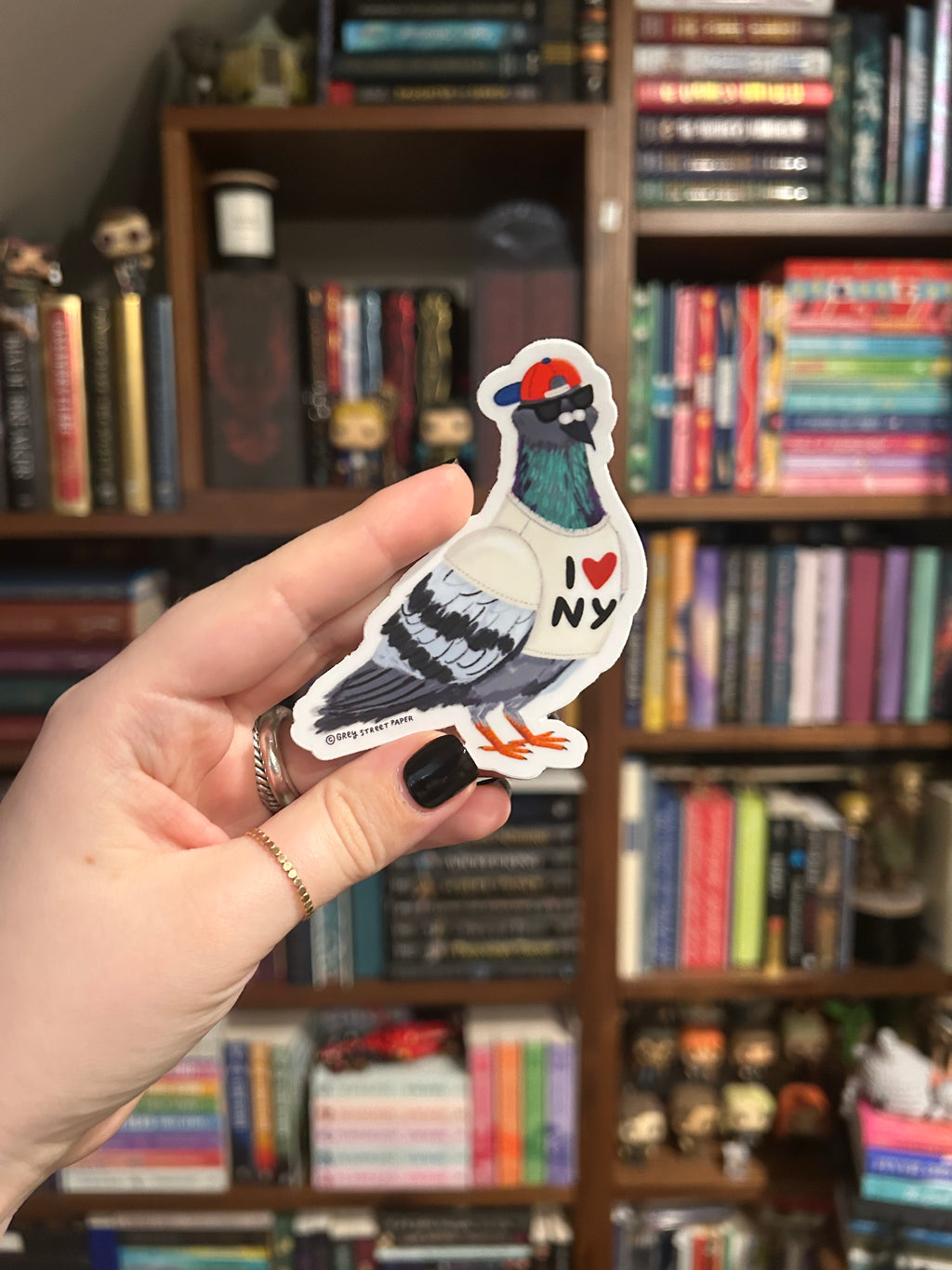 A hand with black nail polish holds a die-cut sticker of a pigeon wearing a red cap and sunglasses, with 'I <heart> NY' written on its chest, against a blurred background of bookshelves.