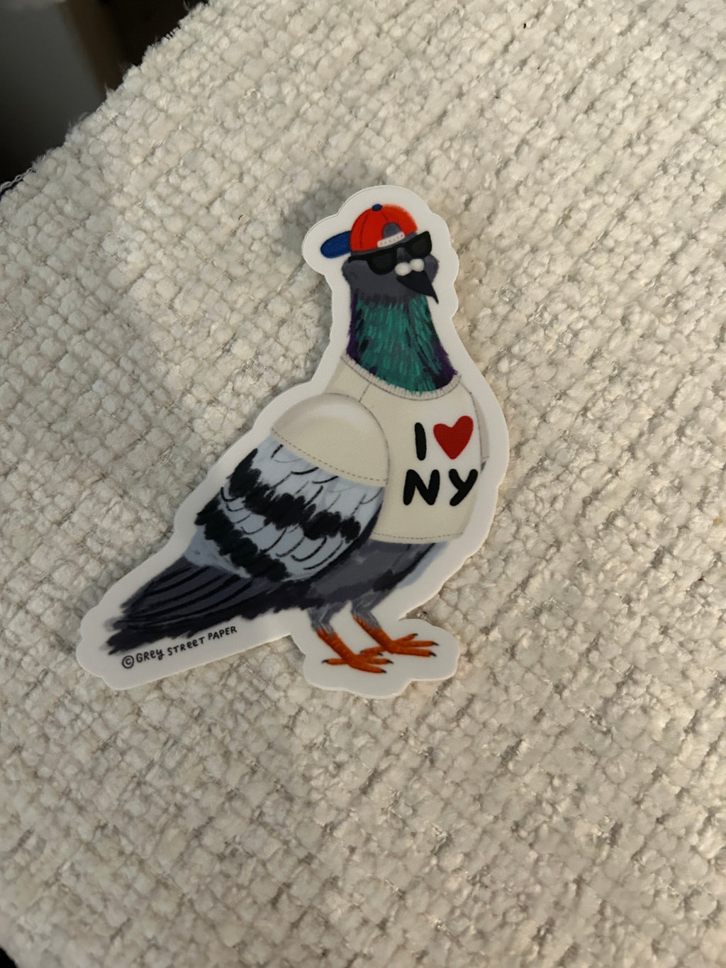 A die-cut sticker of a stylized pigeon wearing a white sweater with an ‘I heart NY' on the chest, placed on a textured off-white fabric background.