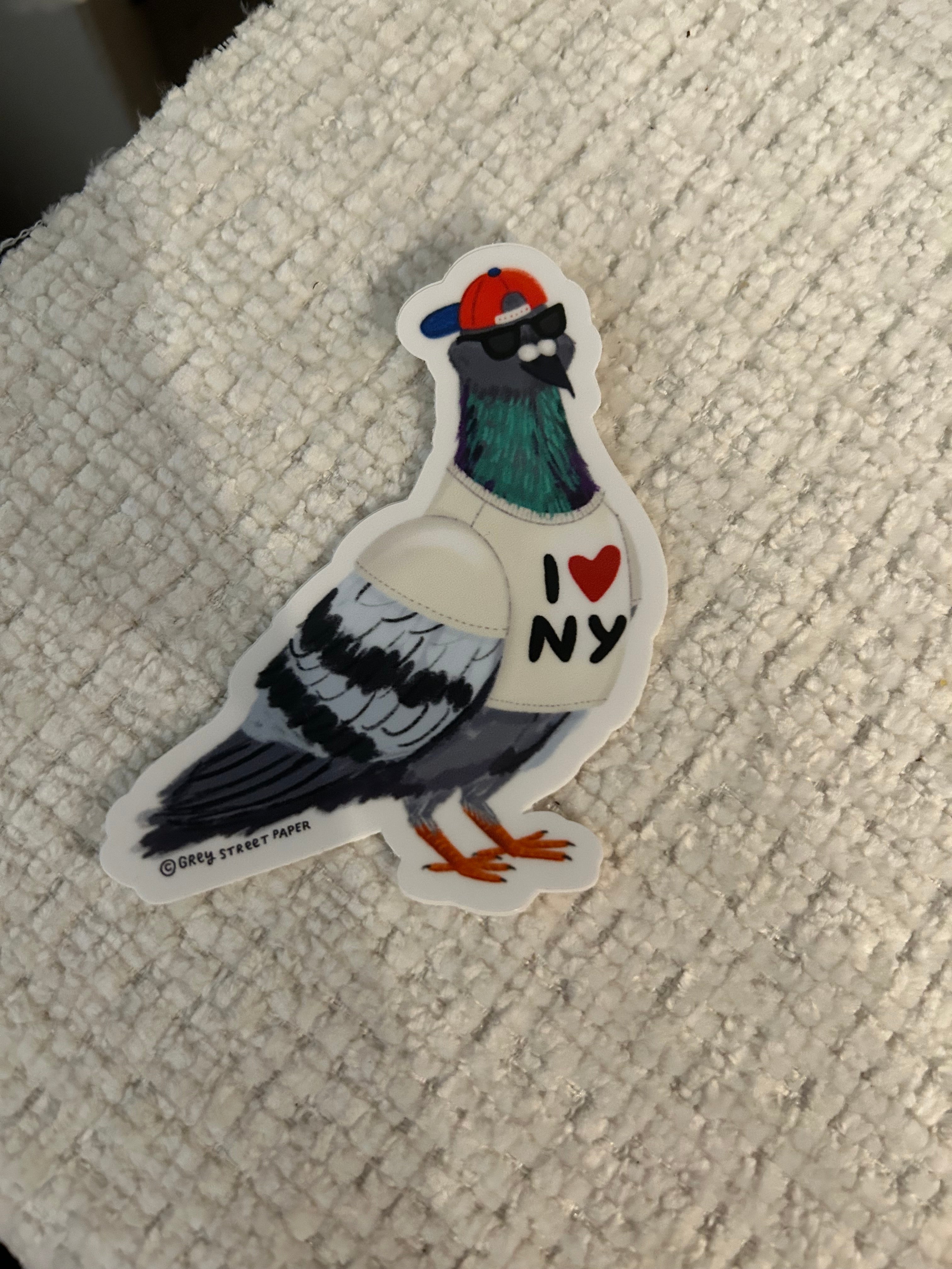 A die-cut sticker of a stylized pigeon wearing a white sweater with an ‘I heart NY' on the chest, placed on a textured off-white fabric background.