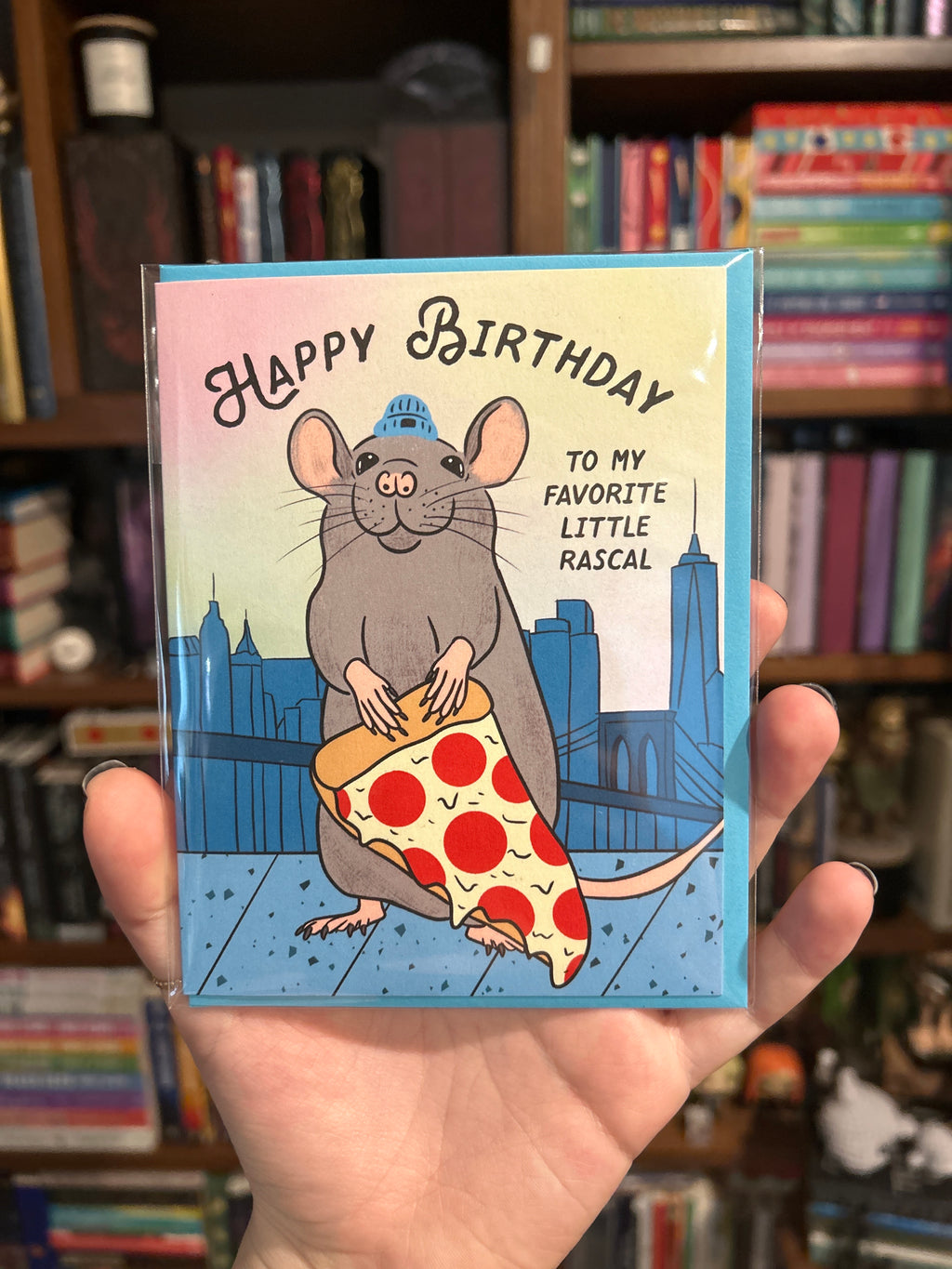 Funny birthday greeting card featuring a smiling New York City rat holding a giant slice of pepperoni pizza with the skyline in the background.