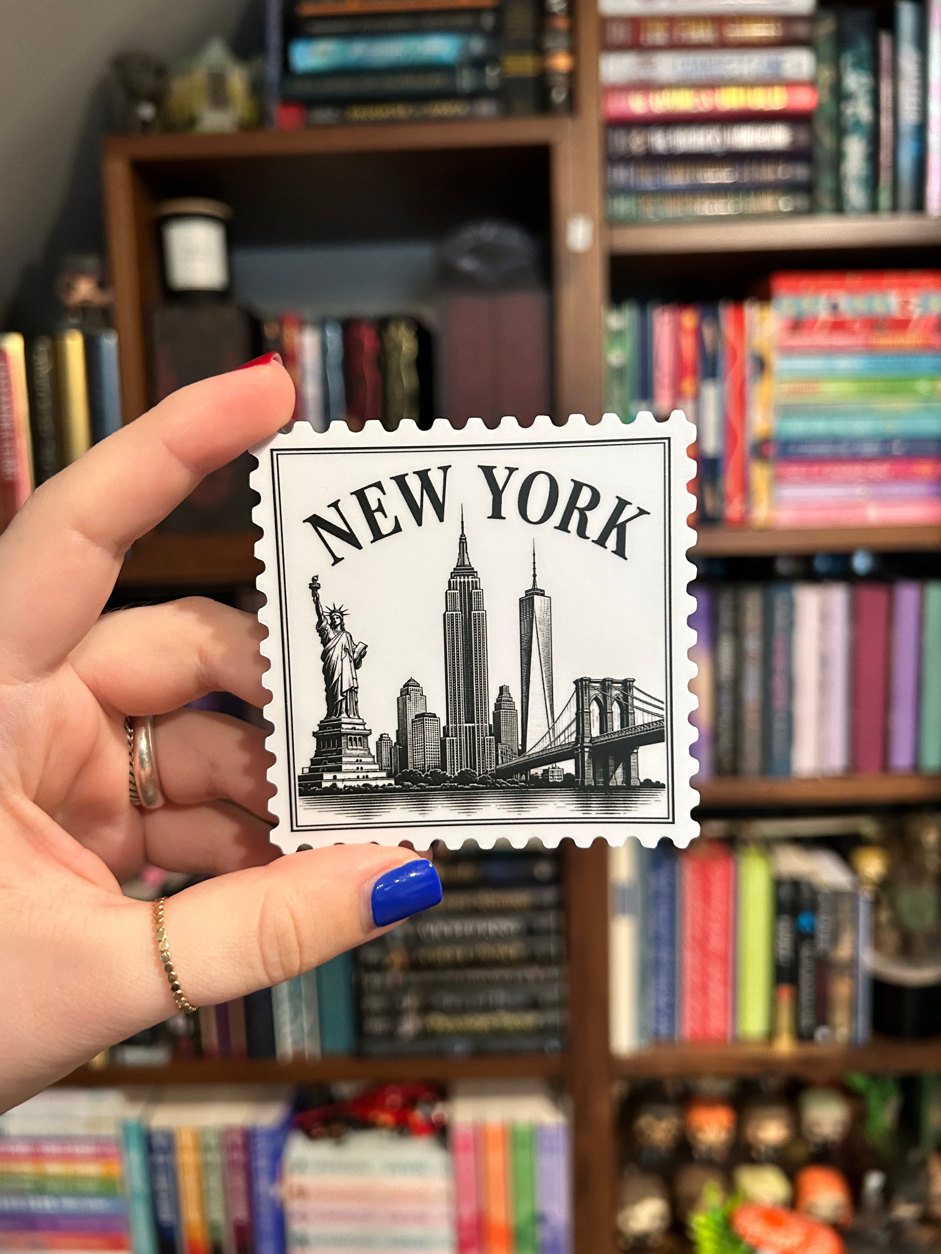 A hand with blue nail polish holds a square white New York City skyline sticker in front of a blurred bookshelf background.