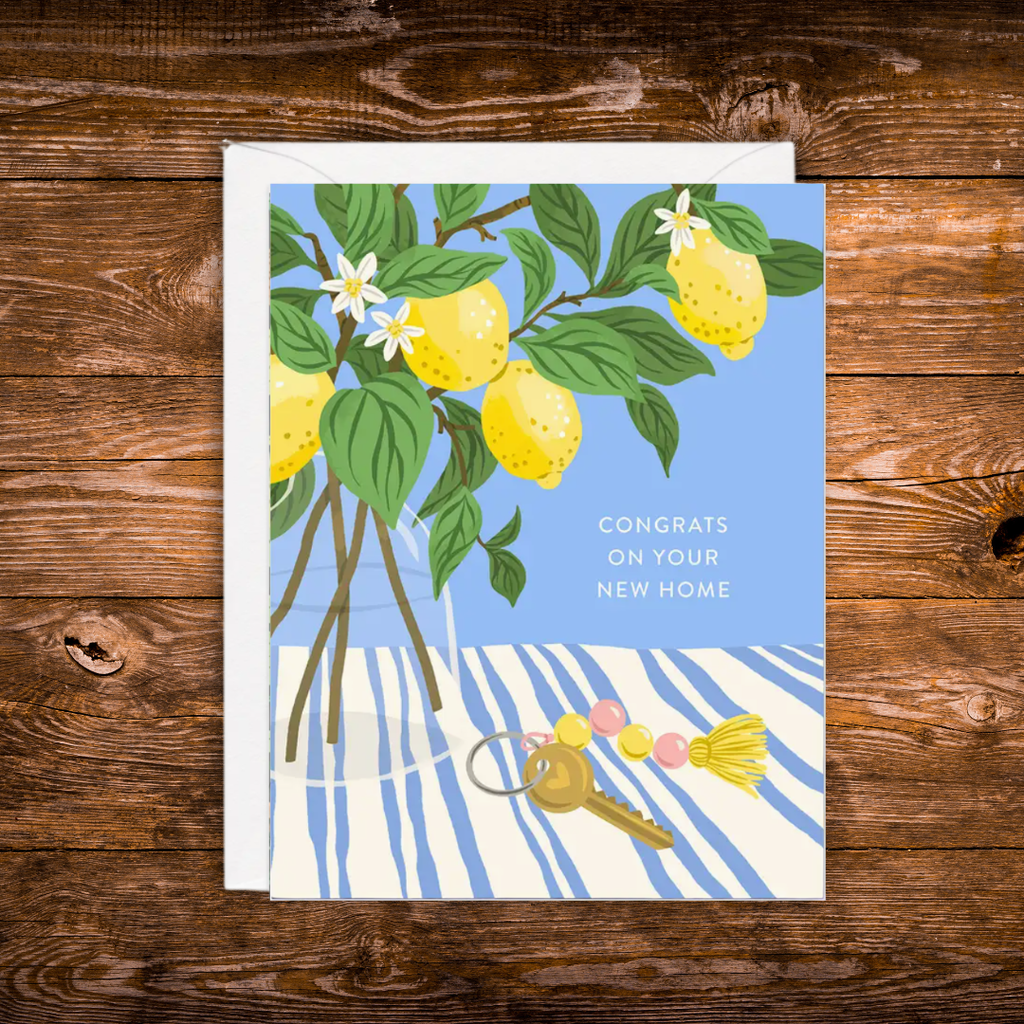 A square greeting card featuring an illustration of lemons on a tree, a glass vase, a striped tablecloth, and keys, with the text 'CONGRATS ON YOUR NEW HOME', photographed on a rustic wooden surface.