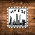 A rectangular New York cityscape stamp-style sticker or print with scalloped edges, featuring black line art of the Statue of Liberty, Empire State Building, One World Trade Center, and Brooklyn Bridge on a white background, placed on a rustic brown wood surface.