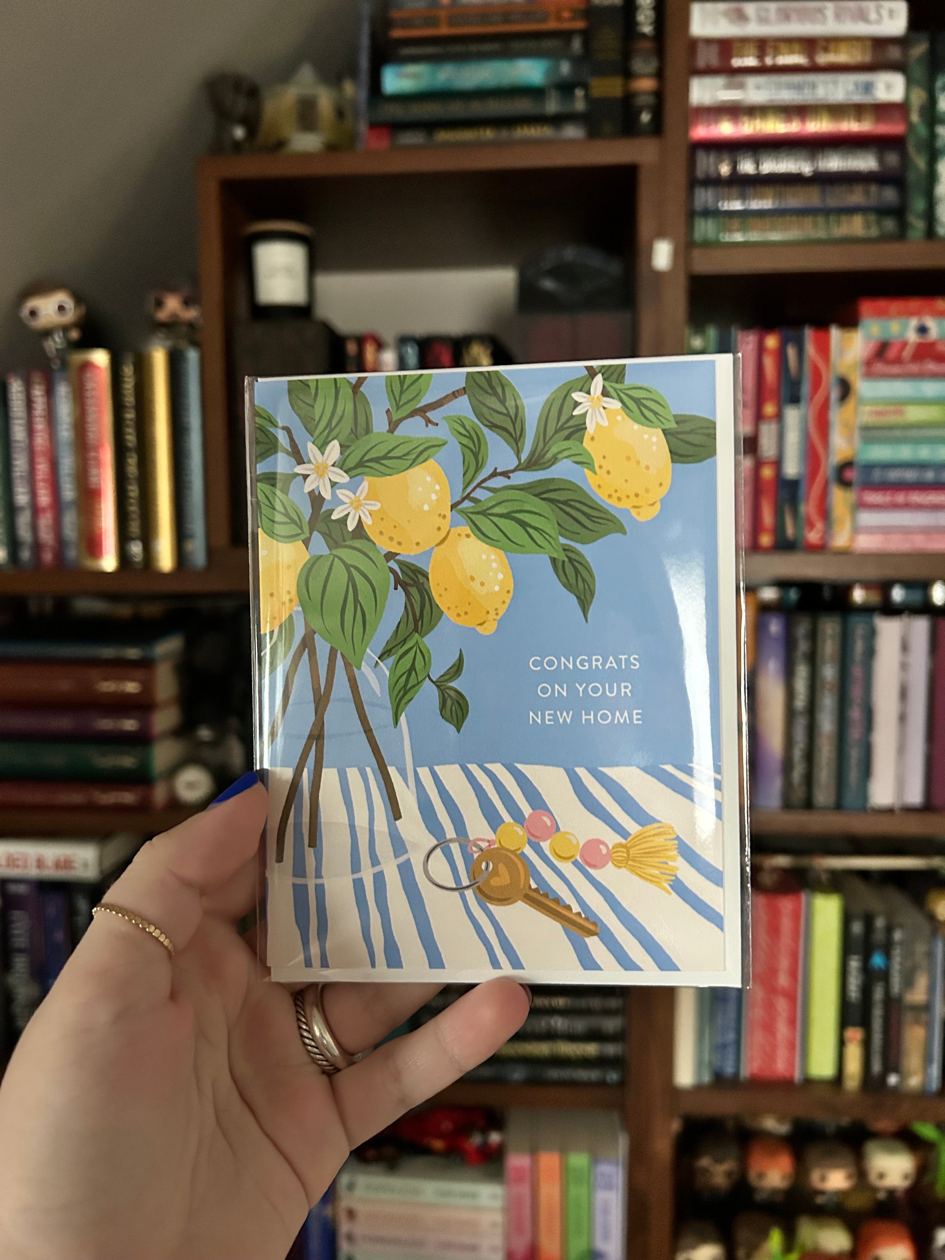 A hand holding a greeting card with a lemon illustration and the text 'CONGRATS ON YOUR NEW HOME' against a blurred bookshelf background.