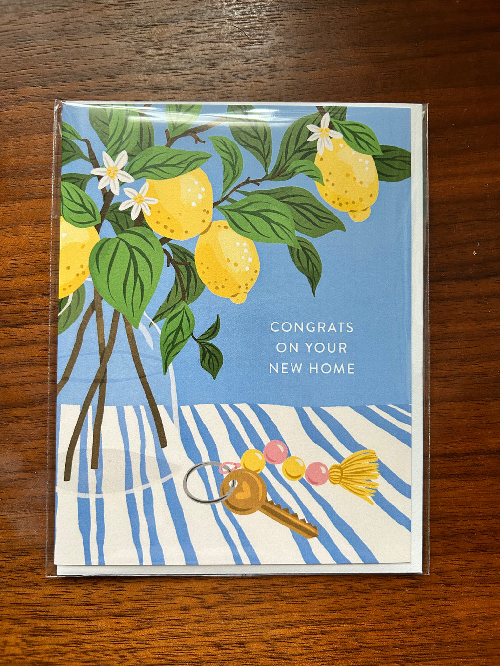 A square greeting card in a clear plastic sleeve, featuring lemons, green leaves, and a key on a blue and white striped background, placed on a wooden surface. The card reads 'CONGRATS ON YOUR NEW HOME'.