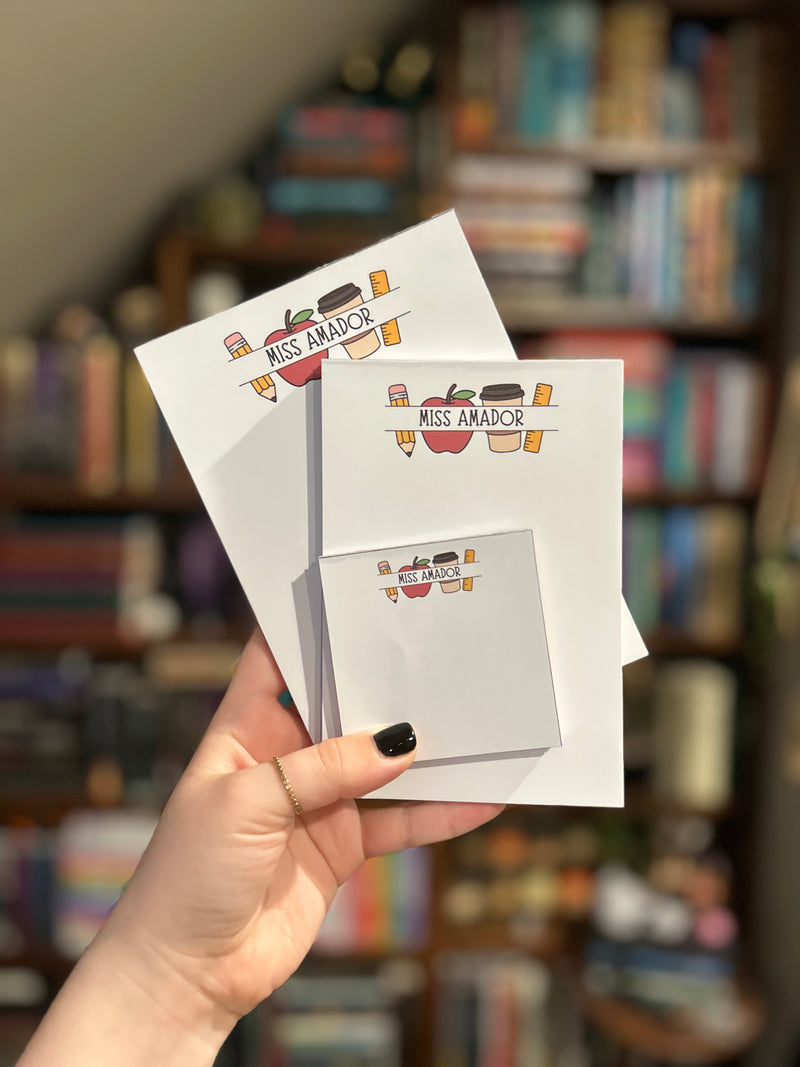 A hand with black nail polish holds three different sized notepads featuring teacher icons: pencil, apple, colorful coffee cup, and ruler illustration and the text 'MISS AMADOR' against a blurred background of bookshelves.