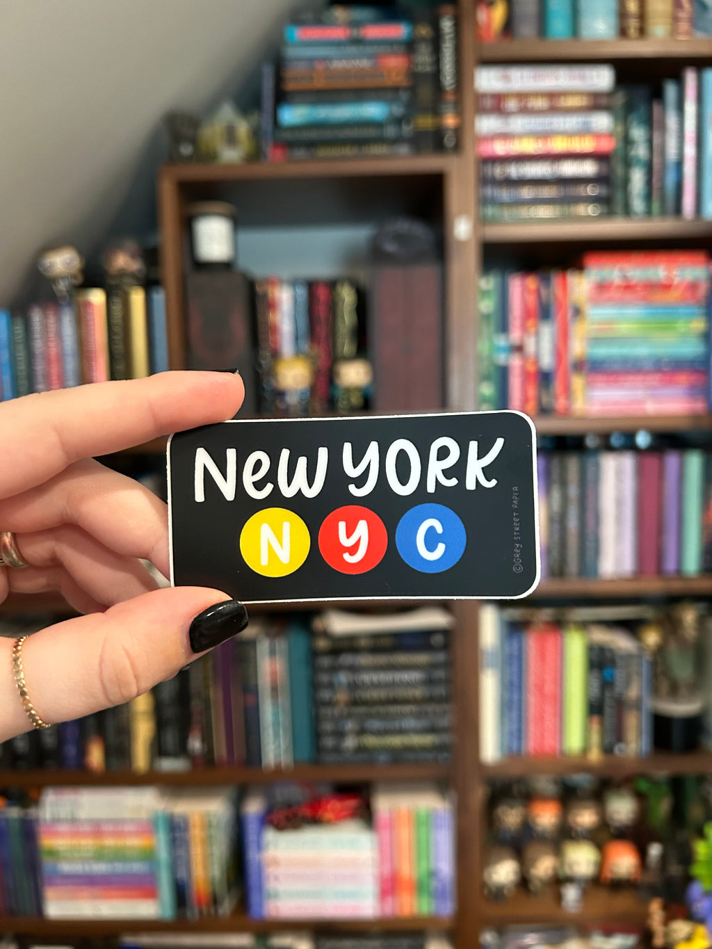 A hand with black nail polish holds a black rectangular sticker featuring the word 'New York' in white alongside blue, red, and yellow circular letters, with blurred bookshelves in the background.