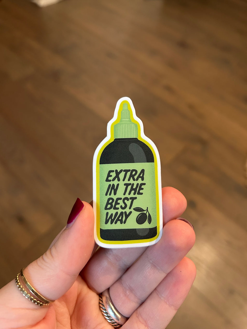 Waterproof vinyl sticker shaped like an olive oil bottle with the phrase "Extra In The Best Way" in green lettering on a light green label.