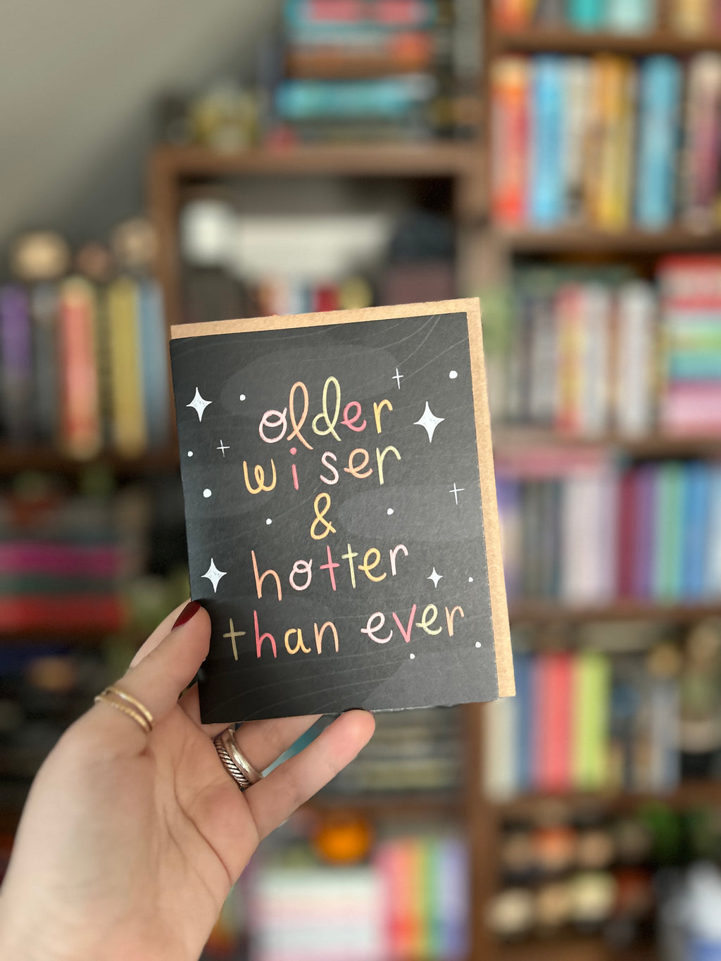Funny birthday greeting card featuring colorful hand-lettered text reading "Older, Wiser & Hotter Than Ever" on a black background with modern celestial accents.