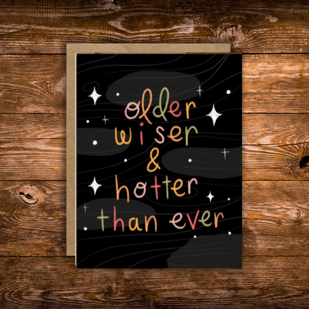 Funny birthday greeting card featuring colorful hand-lettered text reading "Older, Wiser & Hotter Than Ever" on a black background with modern celestial accents.