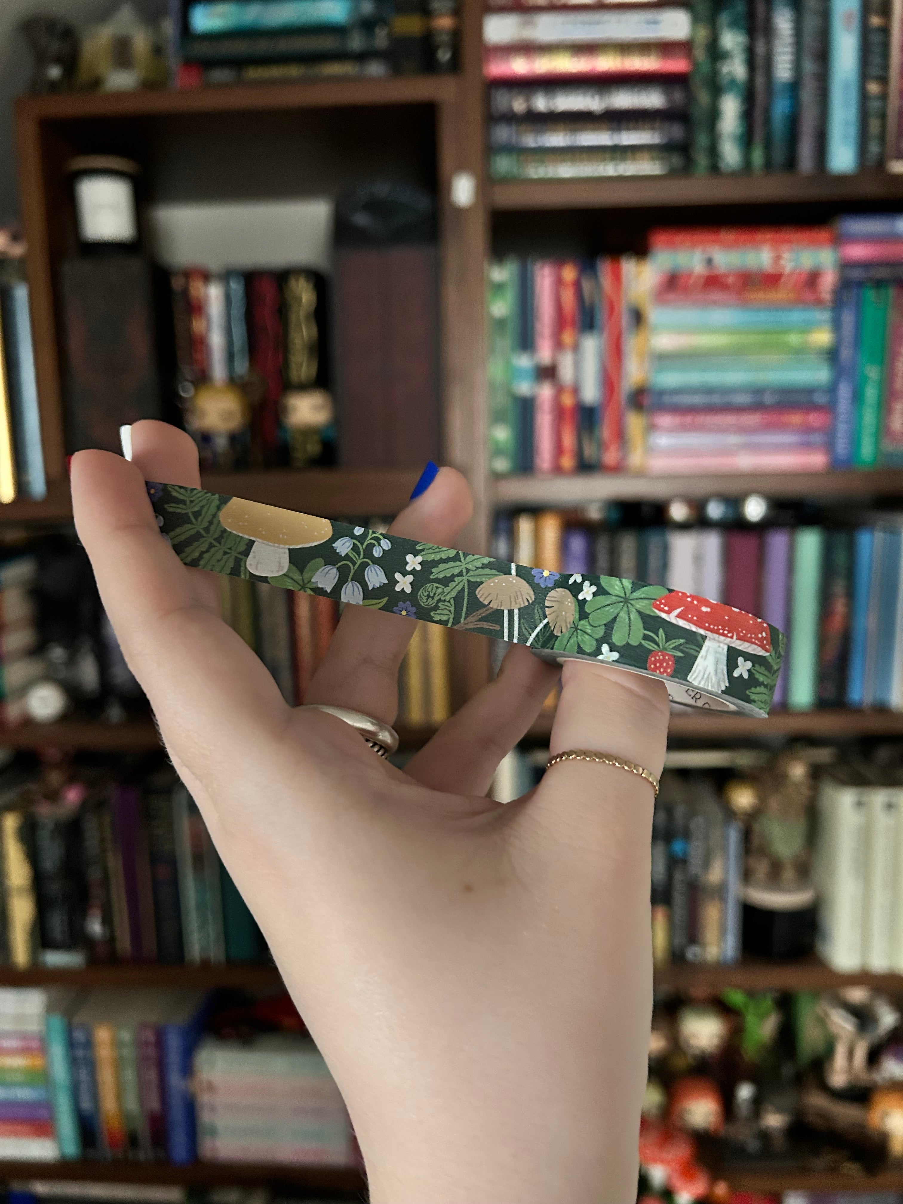 A close-up of a hand holding a slim, patterned wrist strap or lanyard with a green botanical print, set against a blurred background of bookshelves.