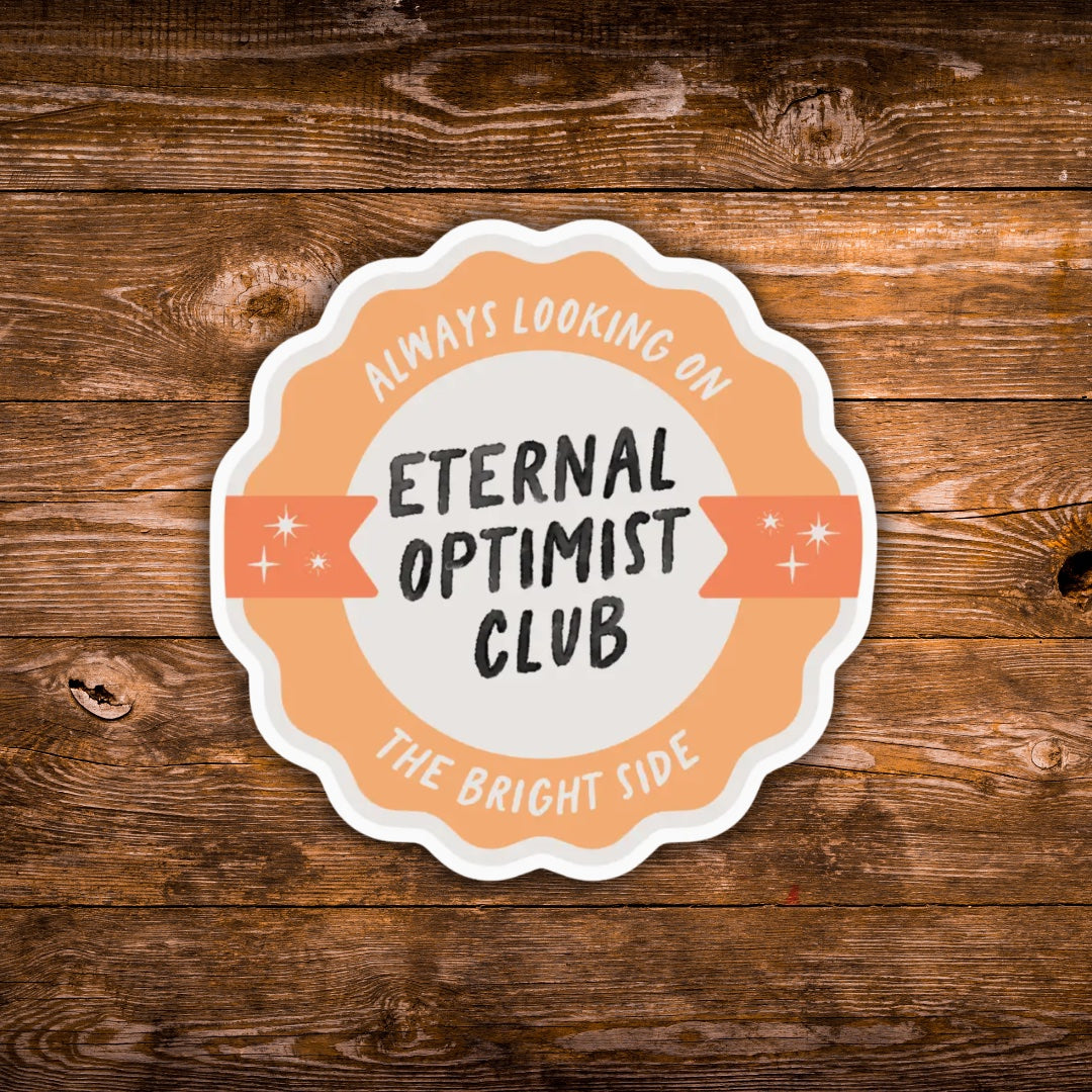 A scalloped die-cut sticker with the text 'ALWAYS LOOKING ON THE BRIGHT SIDE- ETERNAL OPTIMIST CLUB' in black and peach-orange lettering, shown on a rustic brown wooden background.