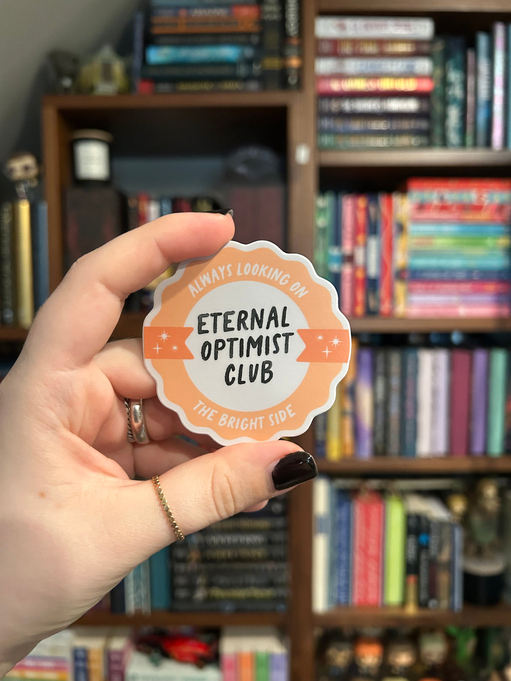 A hand with dark nail polish holds a scalloped die-cut sticker that reads 'ALWAYS LOOKING ON THE BRIGHT SIDE- ETERNAL OPTIMIST CLUB' against a blurred background of bookshelves.