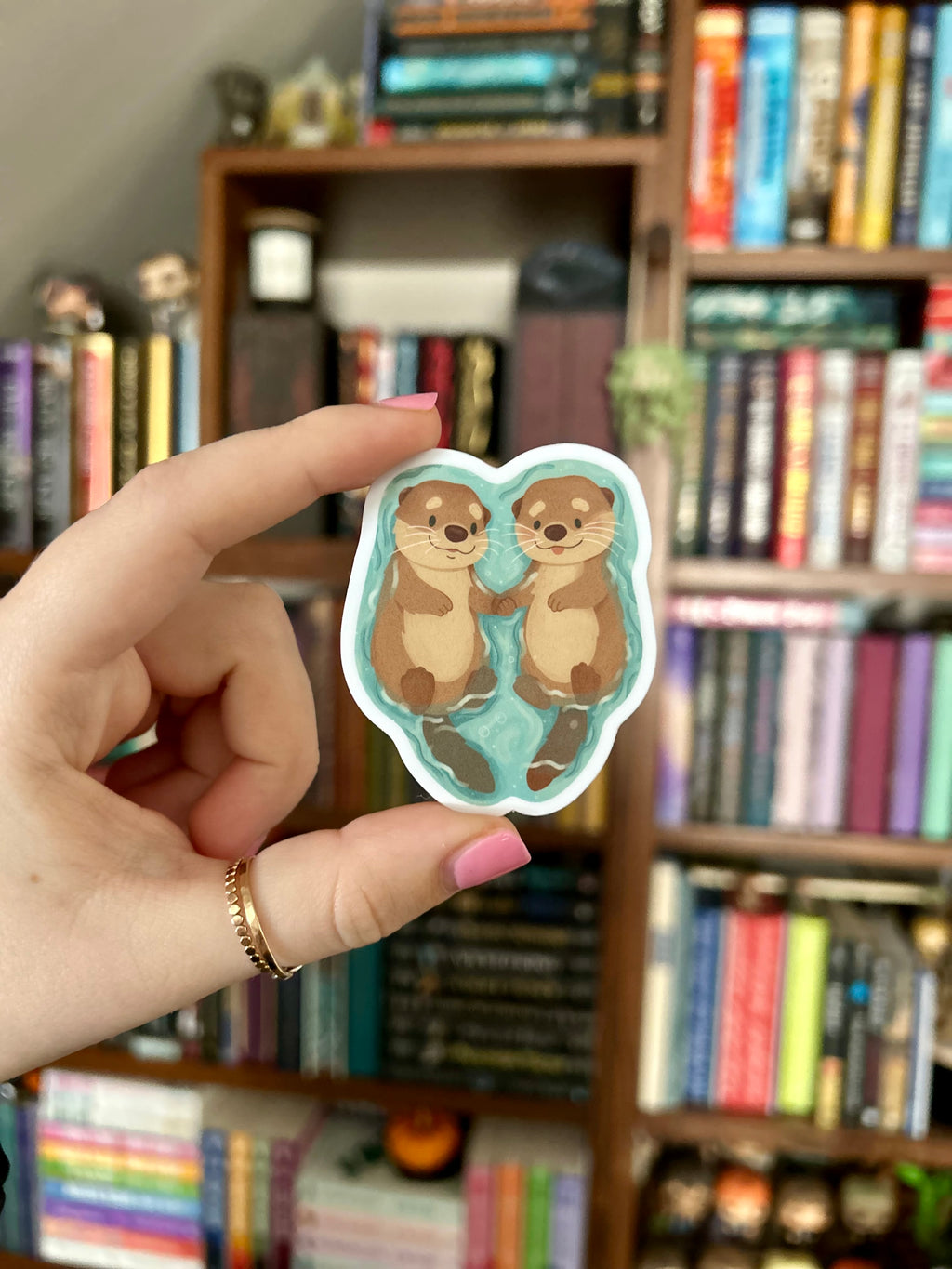 A hand with pink nail polish holds a die-cut sticker featuring two cartoon otters sitting on a light blue background. The sticker is photographed in front of a blurred bookshelf background.