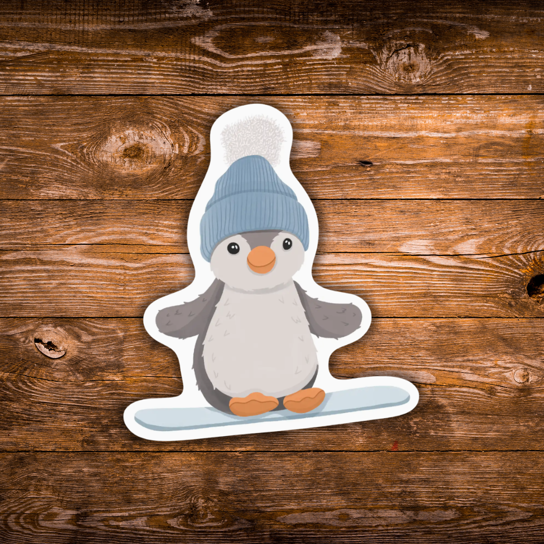 A die-cut sticker featuring a cute cartoon penguin wearing a blue knit hat and standing on a light blue snowboard, shown on a rustic brown wooden background.