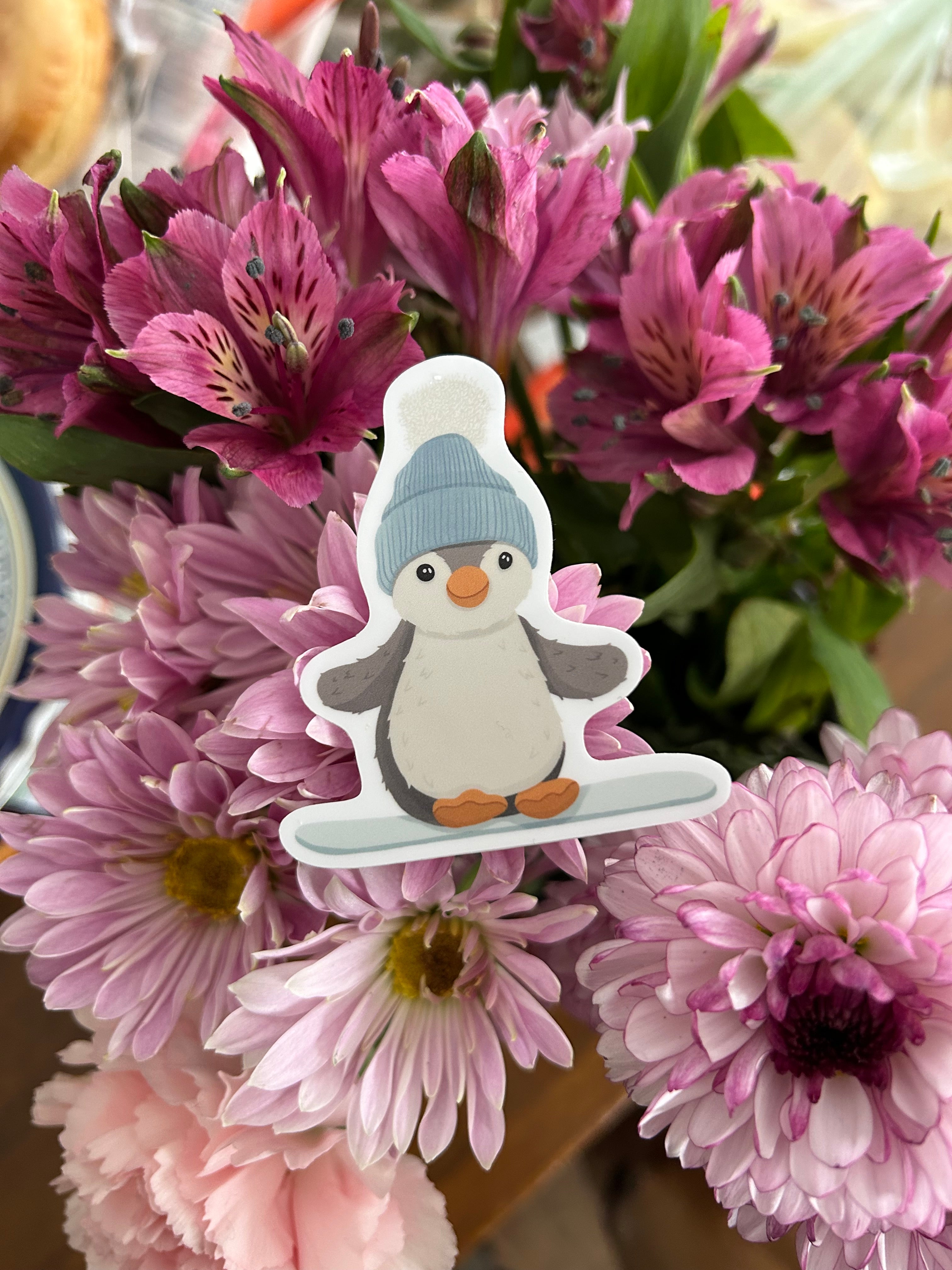A die-cut sticker of a cartoon penguin wearing a blue beanie and holding a snowboard, placed on a bouquet of pink and purple flowers.