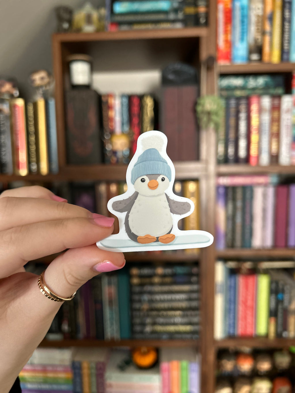 A hand holding a die-cut sticker of a cartoon penguin wearing a blue hat, with a blurred bookshelf in the background.