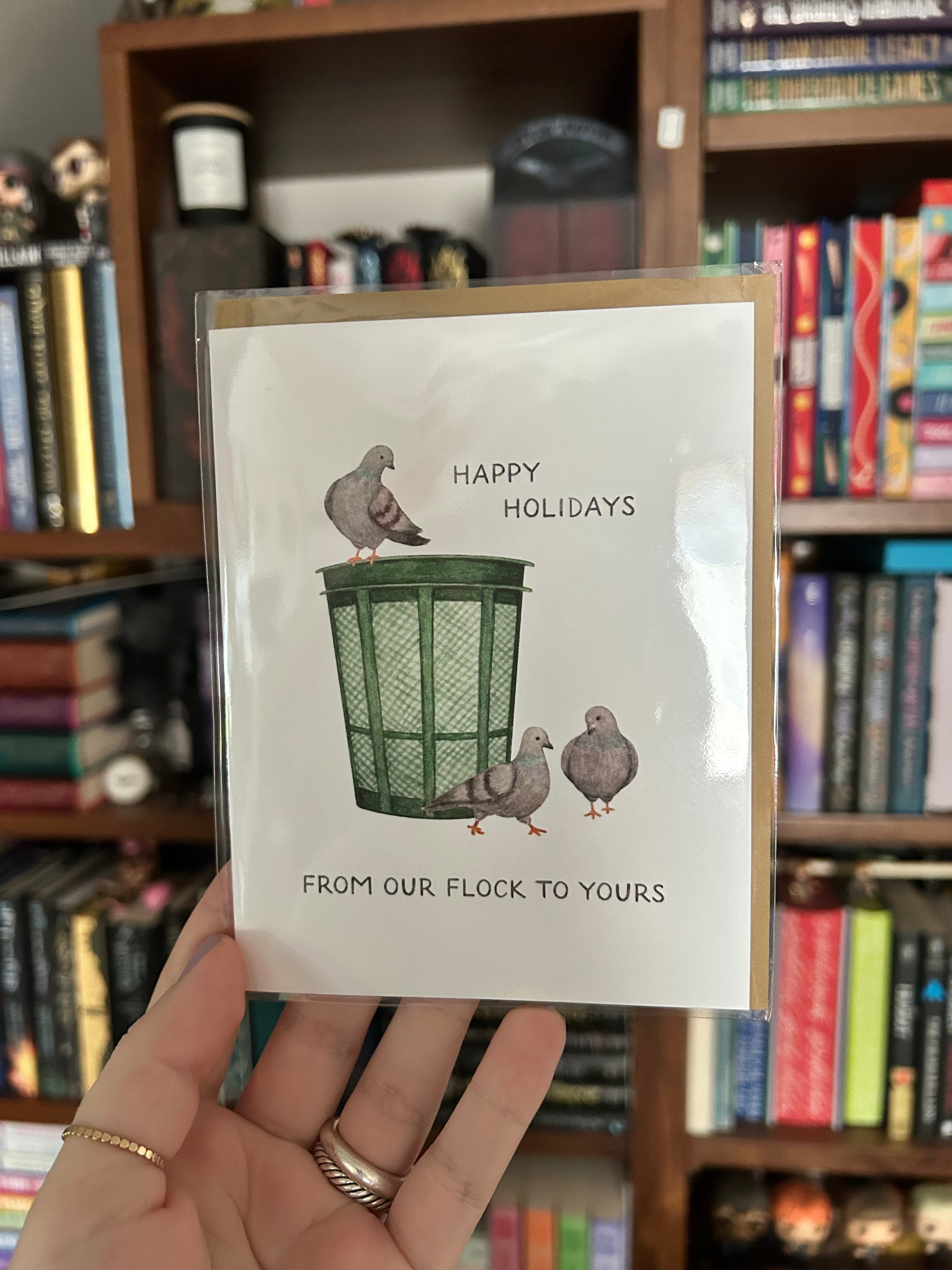 A hand holding a white greeting card with a humorous pigeon-themed holiday design, set against a blurred background of bookshelves.