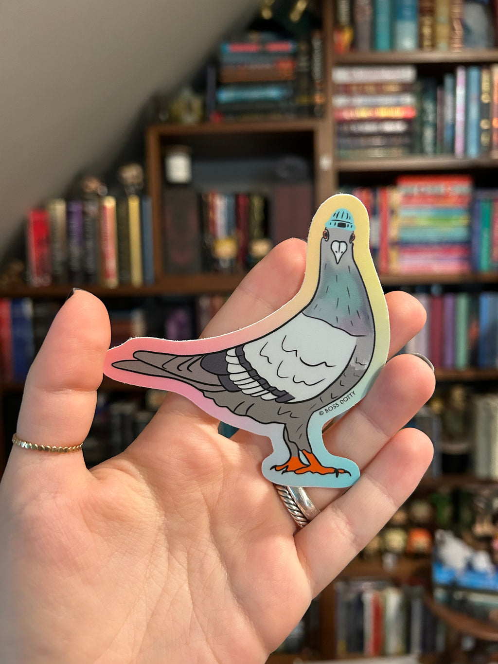 A hand holding a die-cut sticker of a stylized gray pigeon with a white face, blue eye, pink outline, and small orange accent, with blurred bookshelves in the background.
