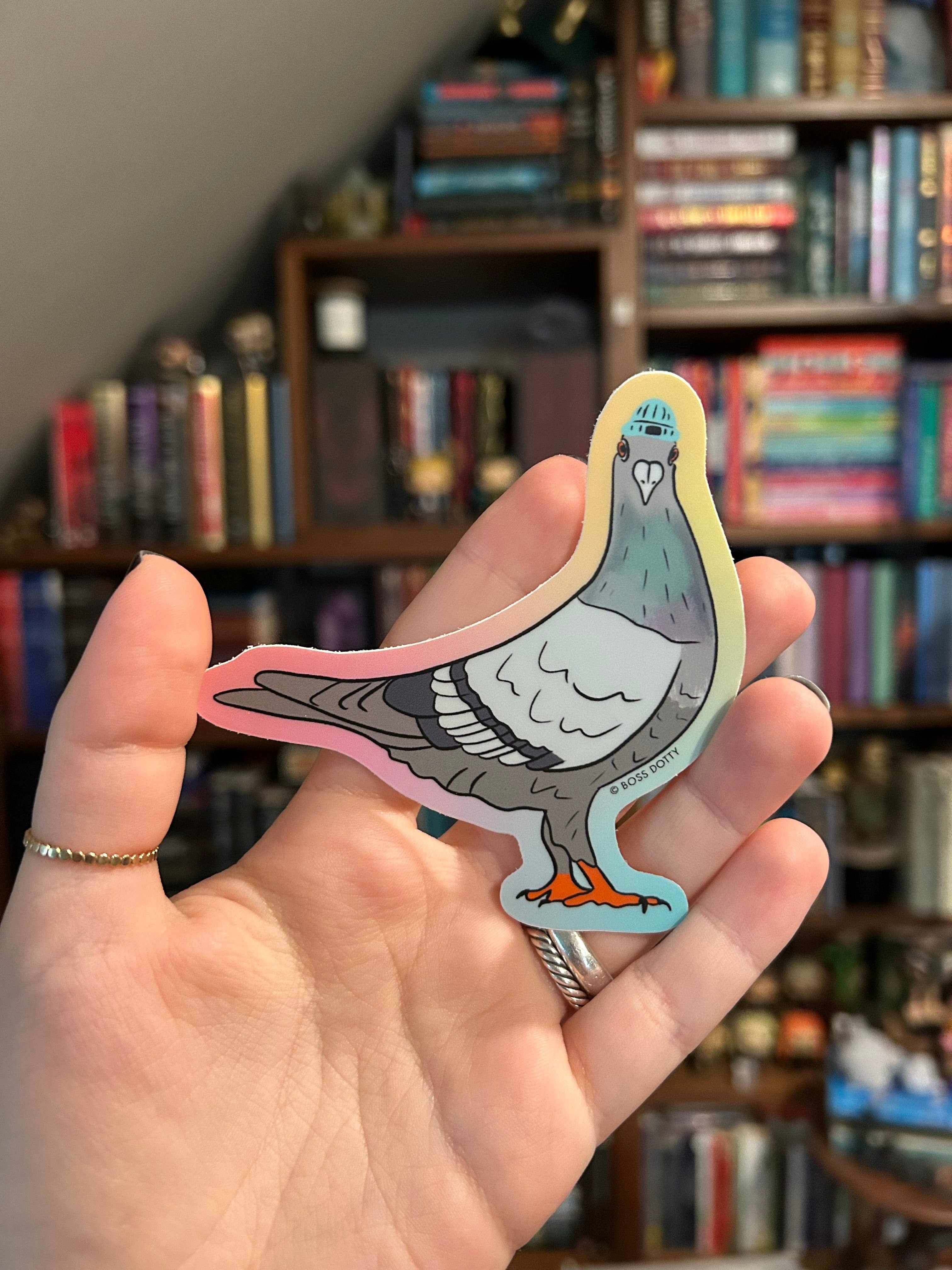 A hand holding a die-cut sticker of a stylized gray pigeon with a white face, blue eye, pink outline, and small orange accent, with blurred bookshelves in the background.