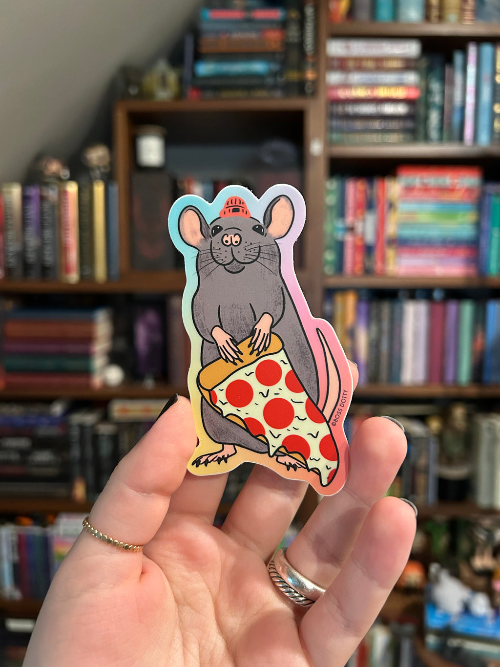 A hand holds a die-cut sticker featuring a cartoon gray rat wearing a small red hat and eating a slice of pepperoni pizza, with a blurred bookshelf background.