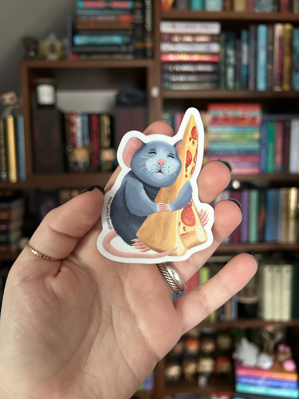 A hand holding a die-cut sticker featuring a blue-gray rat holding a slice of pizza, with a blurred bookshelf in the background.