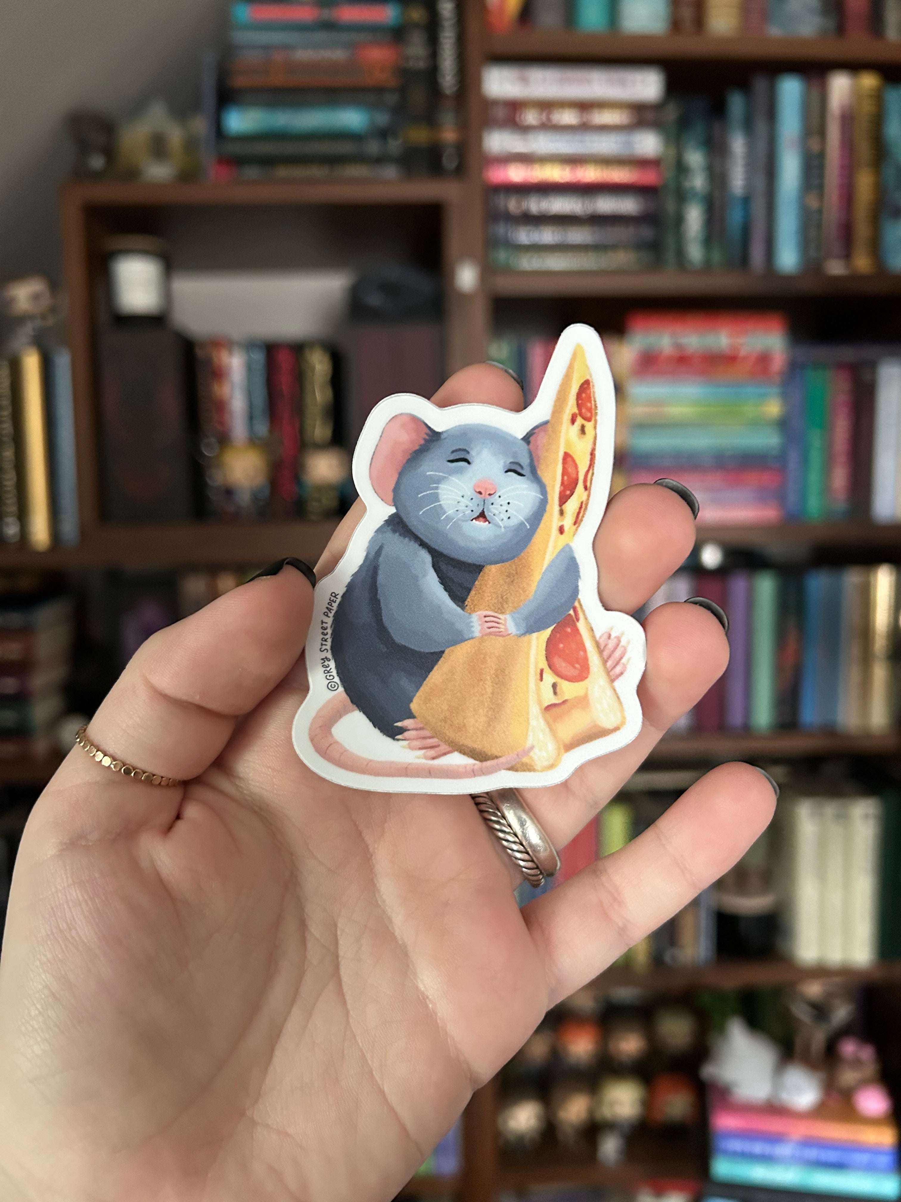A hand holding a die-cut sticker featuring a blue-gray rat holding a slice of pizza, with a blurred bookshelf in the background.
