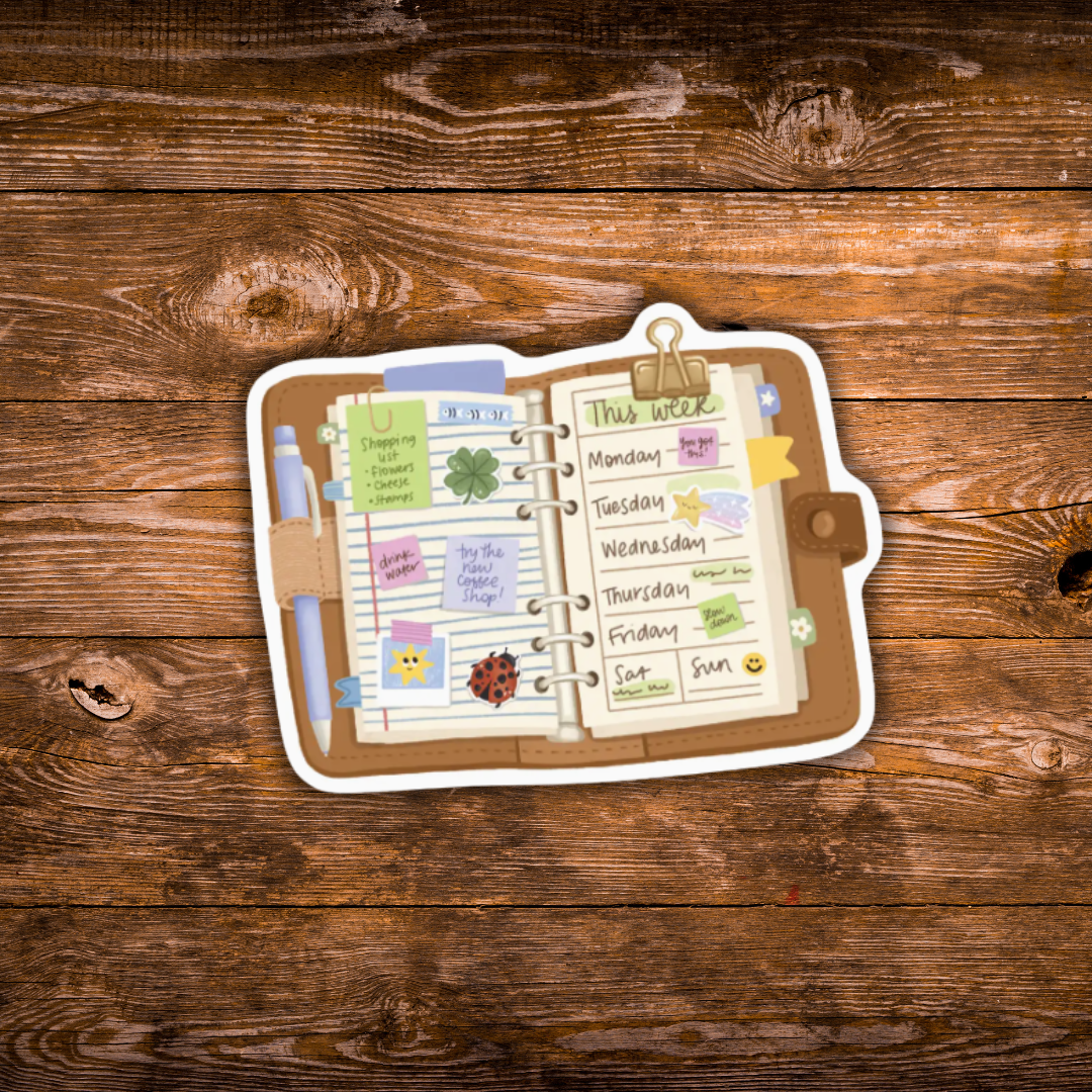 A die-cut sticker depicting an open weekly planner with a pen, sticky notes, and a calendar page, shown on a rustic brown wooden background.