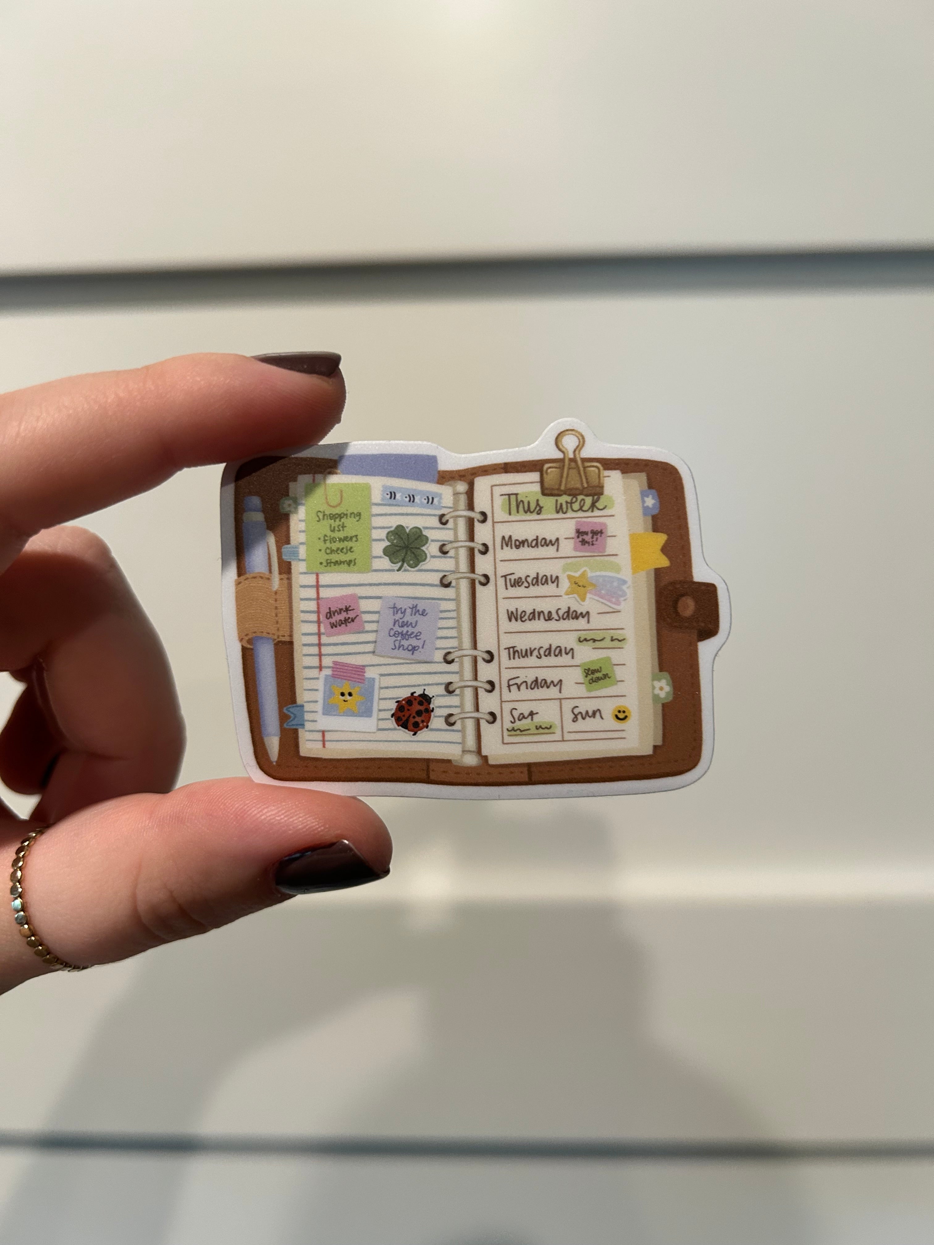 A hand with dark nail polish holds a small illustrated planner or notebook sticker with a brown border and colorful printed pages, against a plain light background.