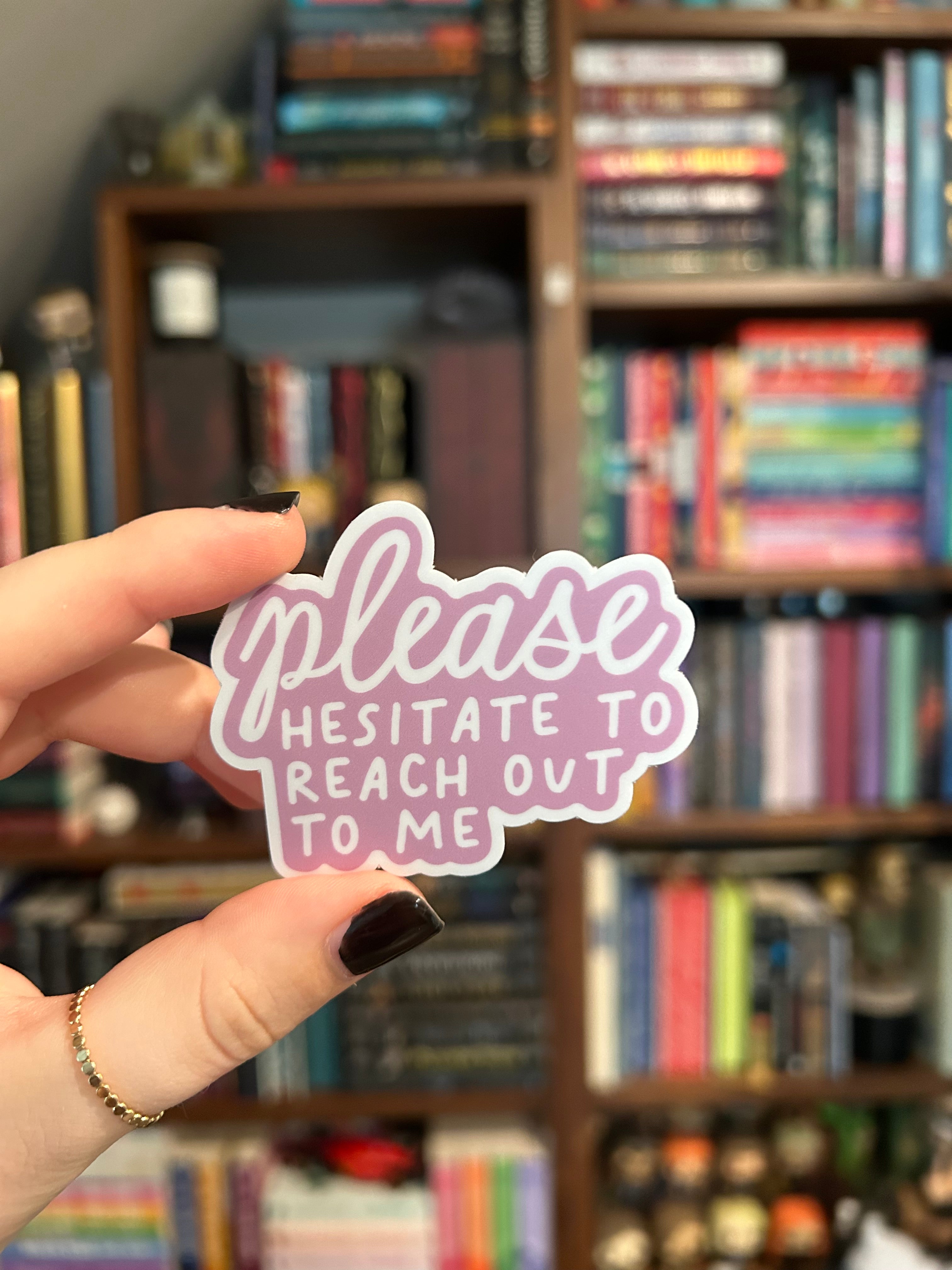 A hand with dark nail polish holds a die-cut sticker in front of a blurred bookshelf. The sticker features the phrase 'please HESITATE TO REACH OUT TO ME' in white script and block lettering with a pink outline.