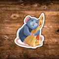 A die-cut sticker featuring an illustration of a blue-gray mouse hugging a slice of pizza, shown on a rustic brown wooden background.
