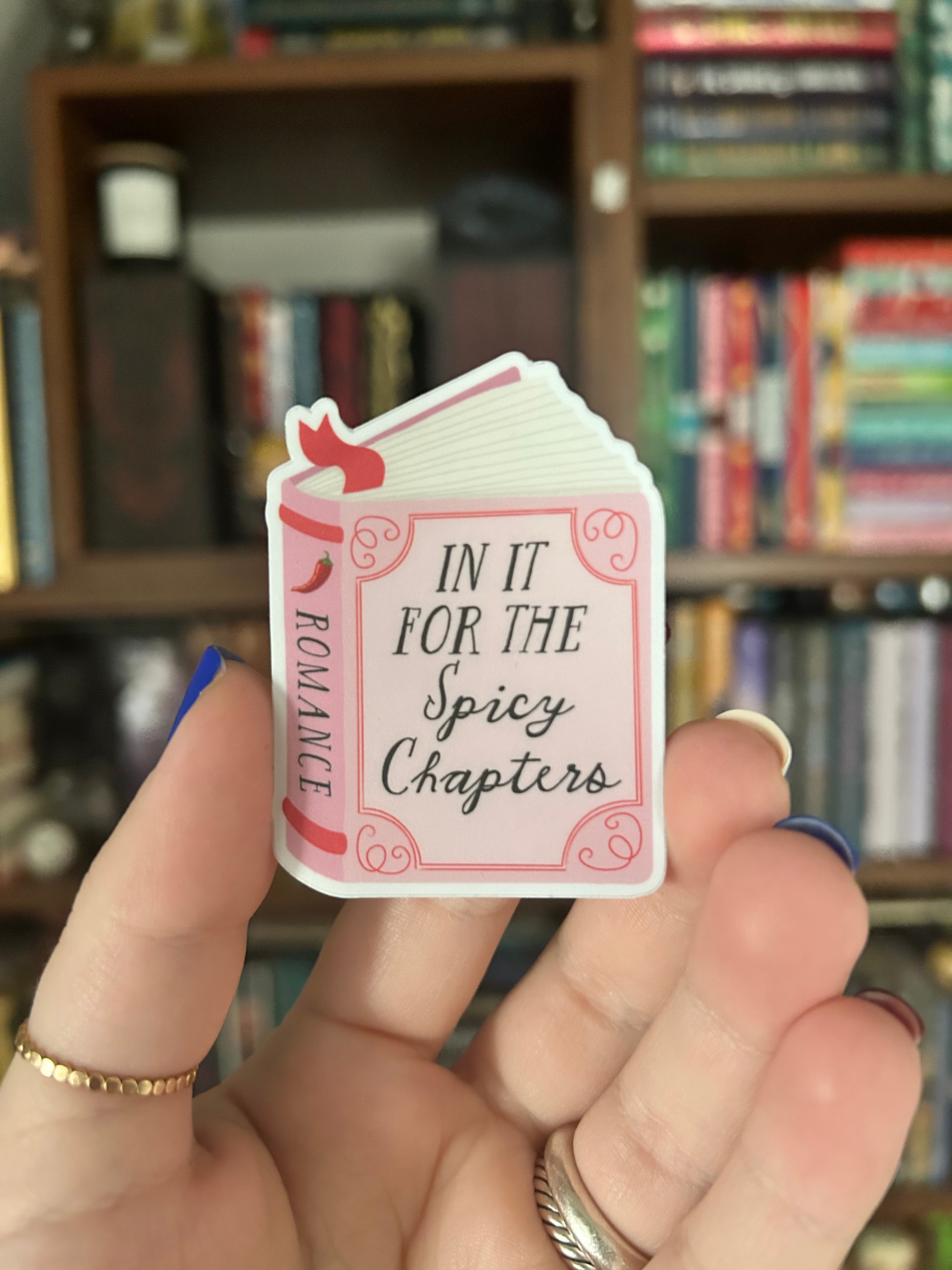 A hand holding a die-cut sticker featuring a pink and white book cover design with the text 'IN IT FOR THE Spicy Chapters ROMANCE'. The background shows a blurred bookshelf.