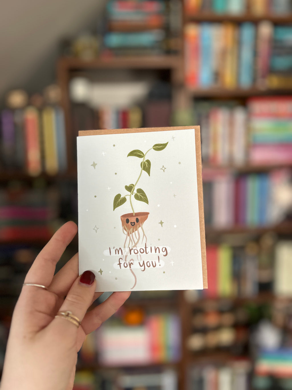 Cute encouragement greeting card featuring a smiling houseplant with exposed roots and the message "I'm Rooting for You!" on a soft blue background.