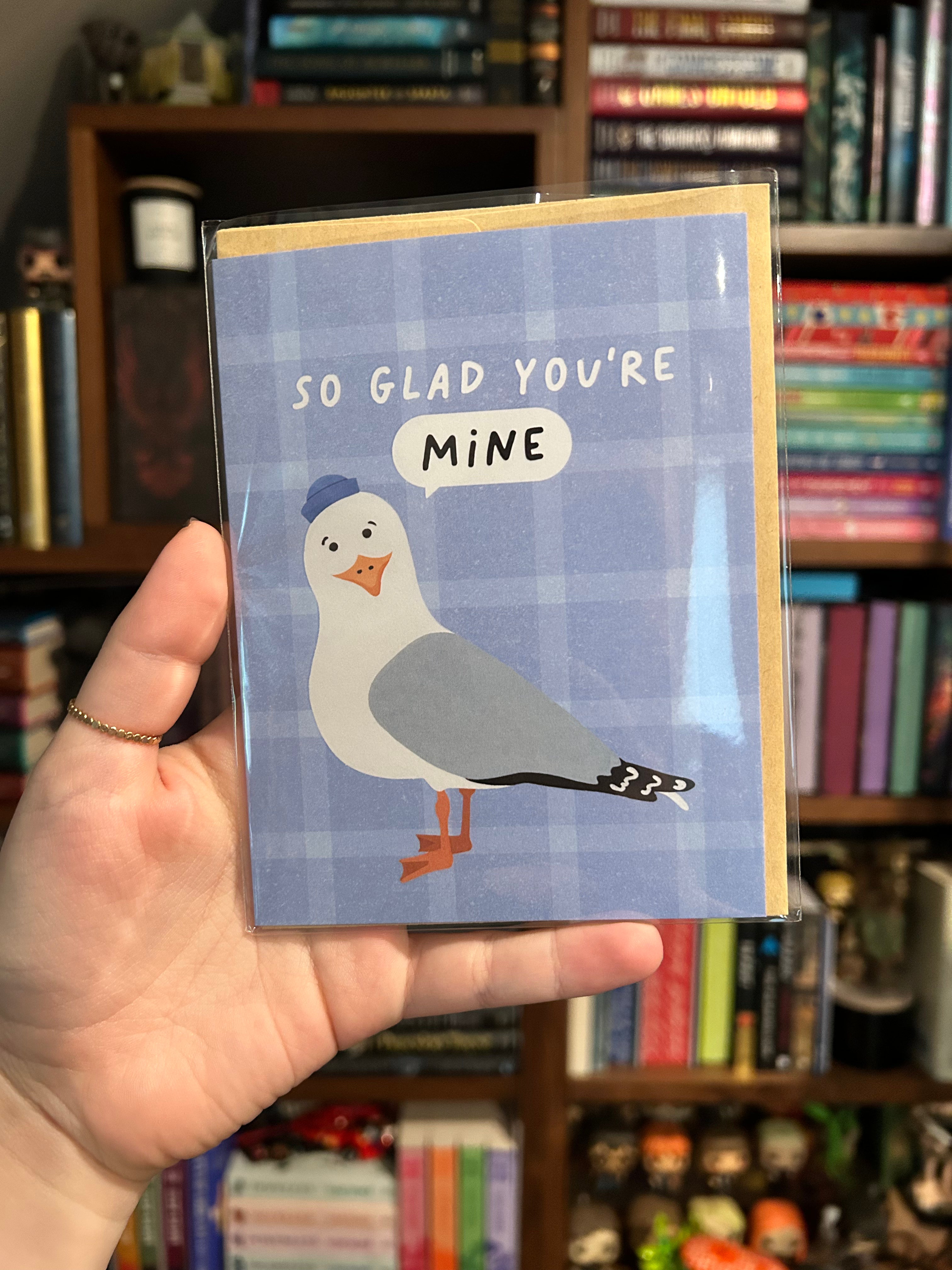 A hand holding a square greeting card with a blue plaid background and an illustration of a seagull with a speech bubble that says 'SO GLAD YOU'RE MINE'. The background shows a blurred bookshelf.