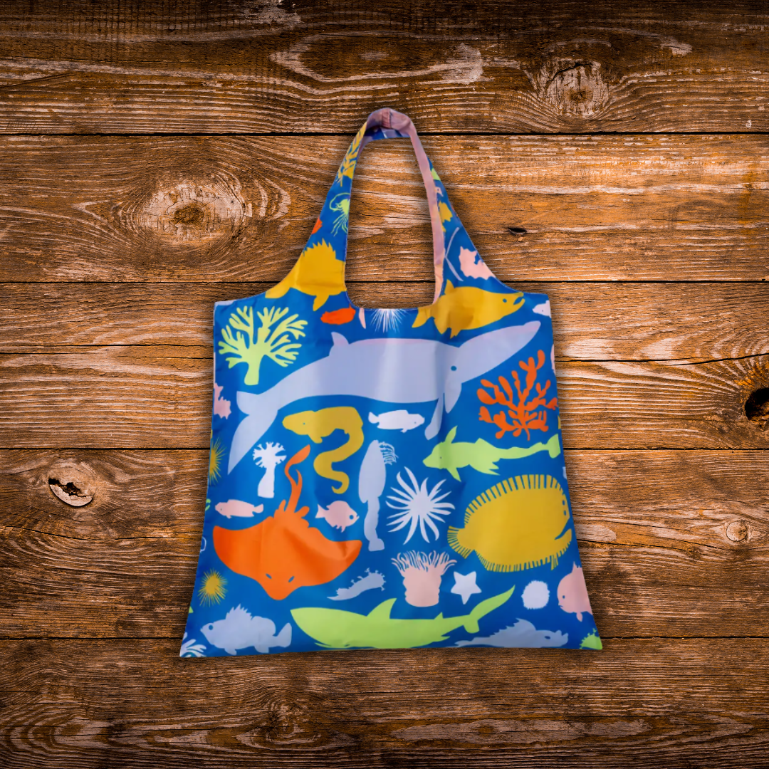 A reusable tote bag with an ocean-themed multicolor print, featuring illustrations of whales, fish, coral, and sea life on a blue background, displayed flat on a rustic wooden plank surface.