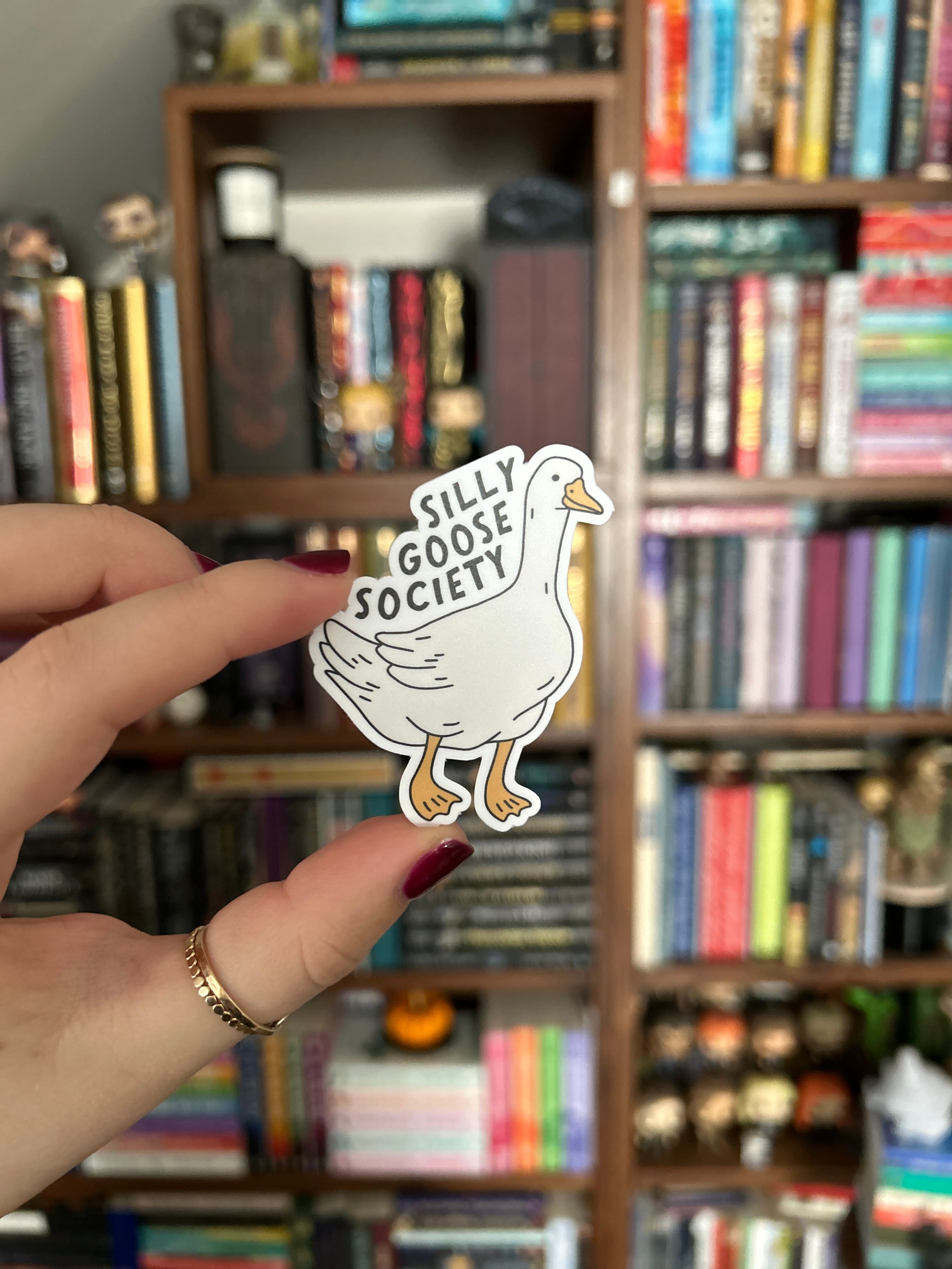 A hand holds a die-cut sticker featuring a white goose with black text reading 'SILLY GOOSE SOCIETY' against a blurred background of bookshelves.